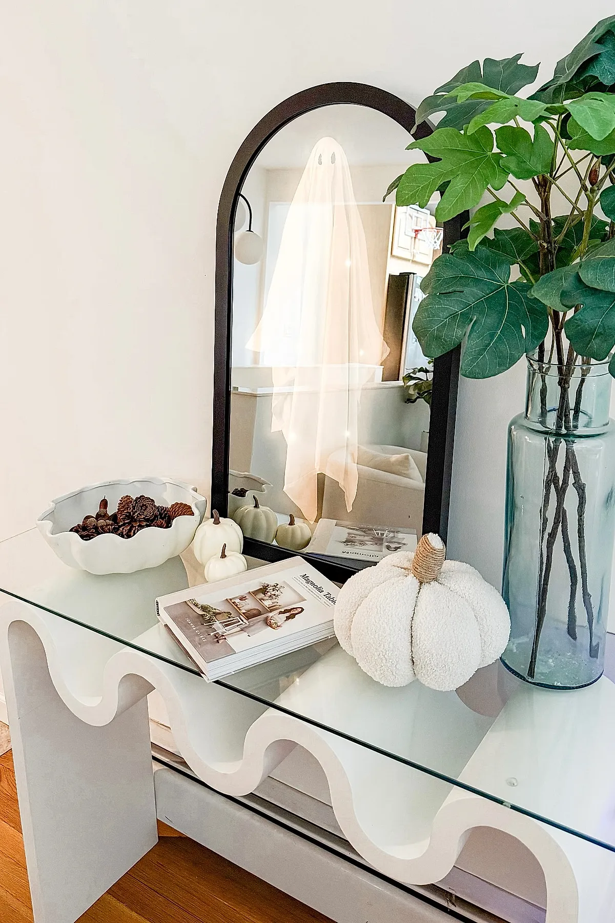 Modern fall decor featuring a black arched mirror with a sheer ghost silhouette reflection, white textured pumpkins, pinecones in a white bowl, a book on a wavy white glass-top table, and green leaves in a tall clear blue-green glass vase.