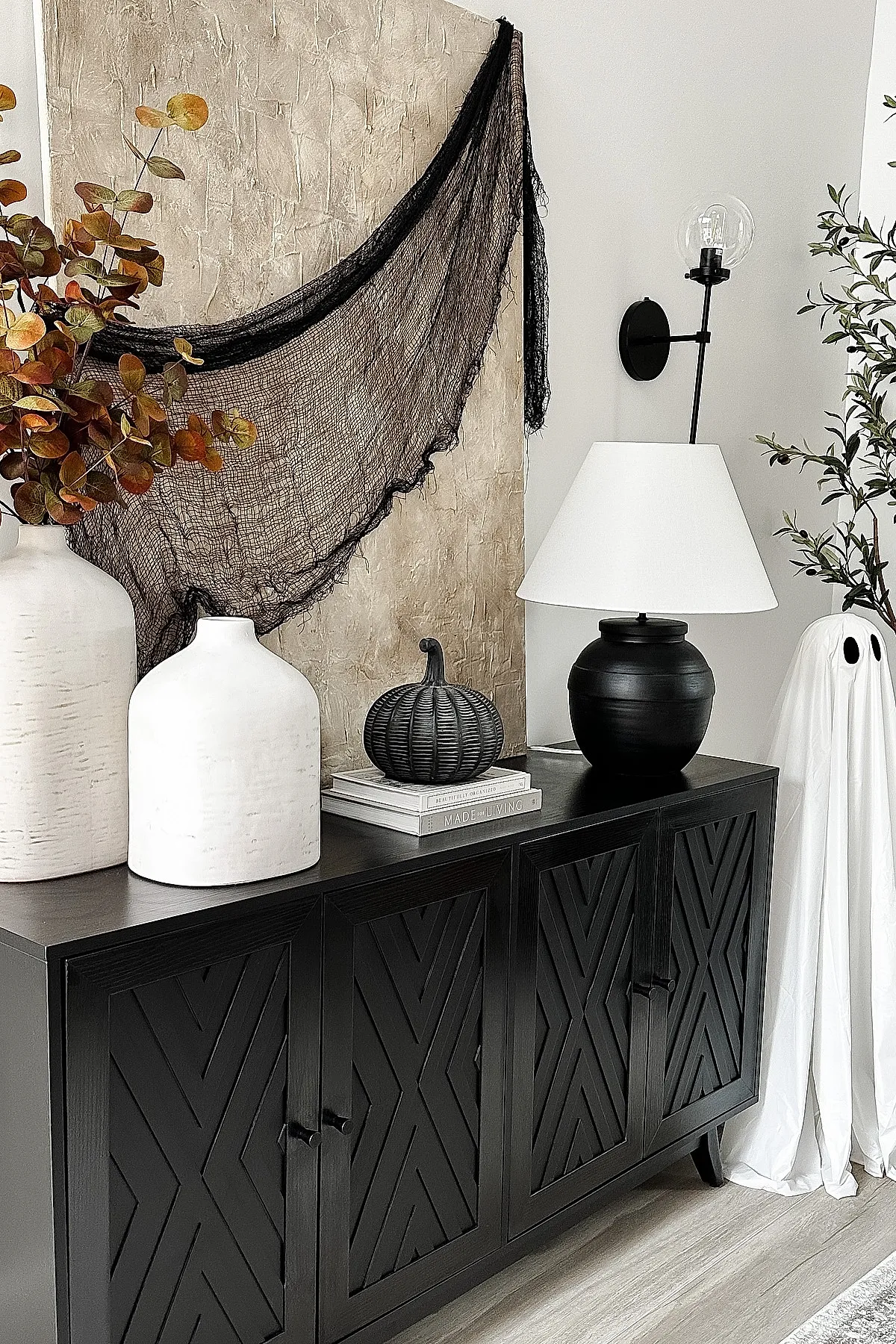 Black geometric console with large white textured ceramic vases holding dried eucalyptus, a matte black pumpkin on stacked books, sheer black fabric draped over textured beige wall, black lamp with white shade, and a minimalist white ghost decoration.