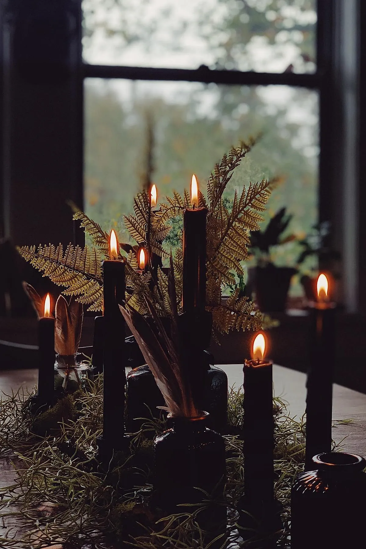 Black taper candles burning on a wood table surrounded by dried golden fern leaves, green moss-like foliage, and small glass jars holding brown feathers.