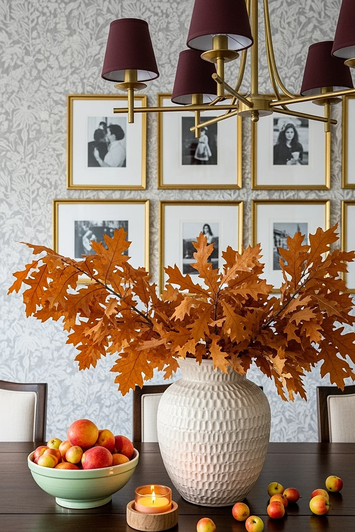Large white textured ceramic vase filled with bright orange oak branches on dark wood table next to a green bowl of red and yellow apples, scattered small apples, and a lit candle in wooden holder, set against floral wallpaper with gold framed black-and-white photographs.