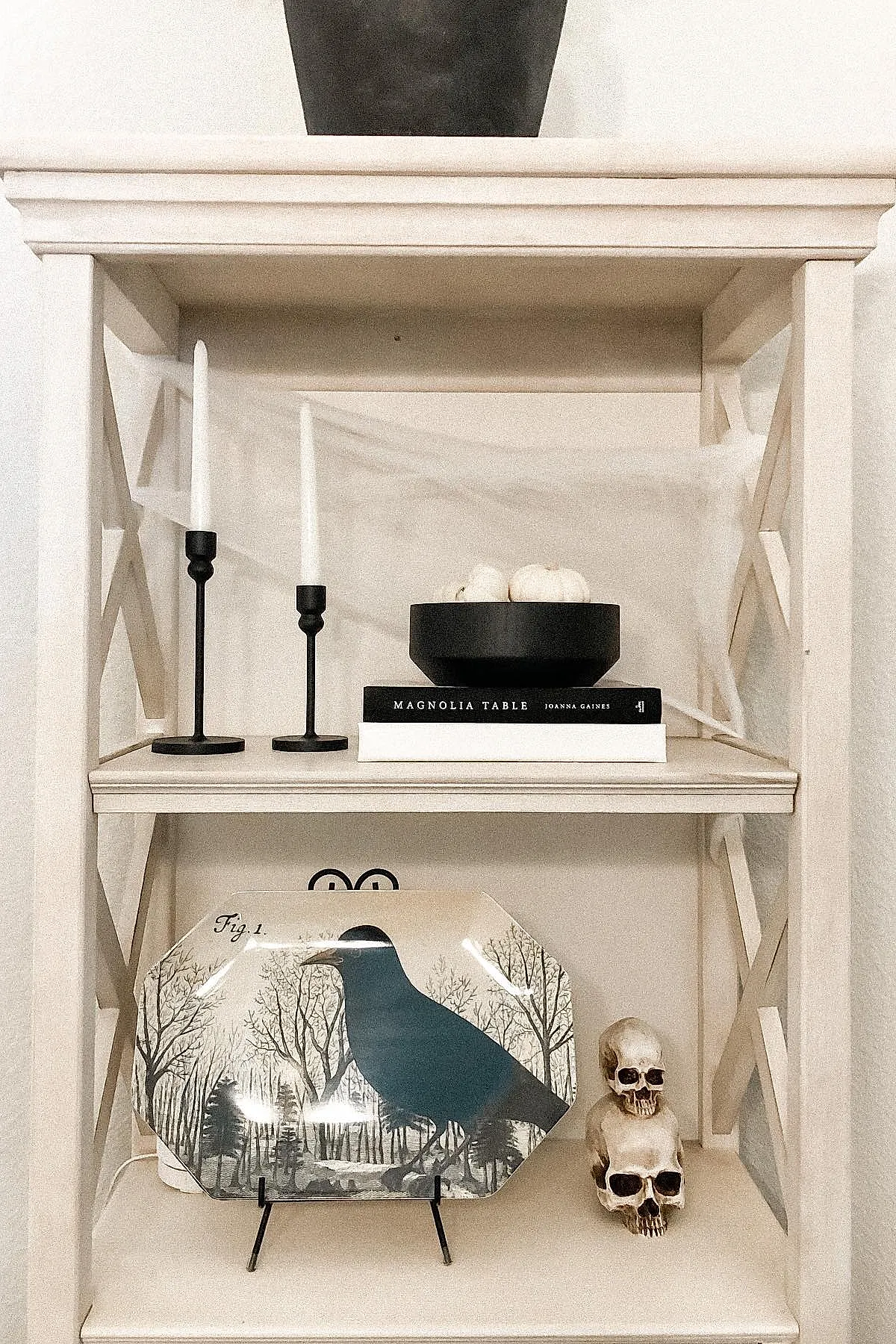 Light cream shelf decorated with a large rectangular plate featuring a black raven against bare trees, two stacked realistic skull decorations, two black candle holders with white taper candles, and a stack of books topped by a black bowl holding small white pumpkins.
