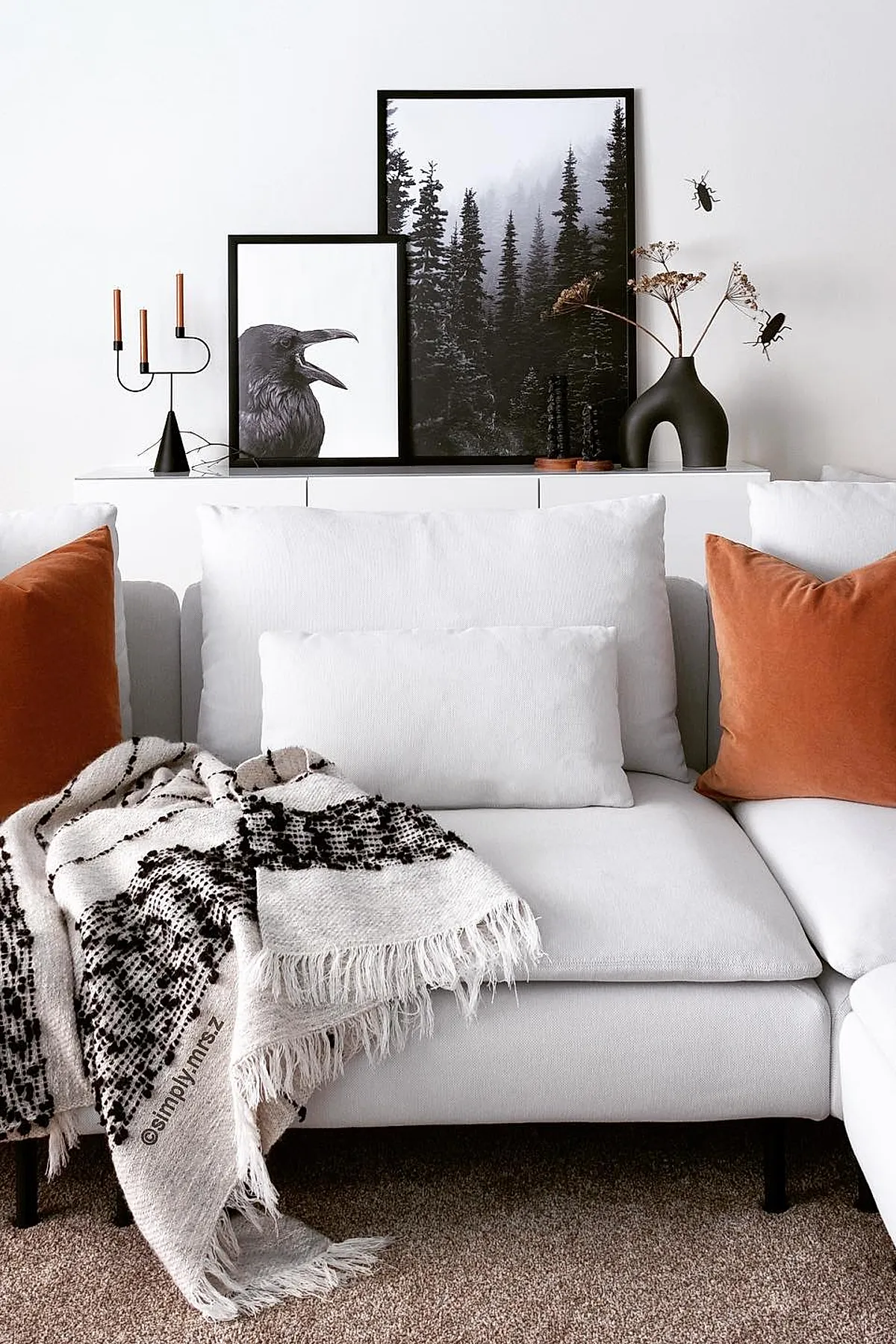 White sofa with rust orange pillows and cream throw blanket in front of white cabinet topped with two black-framed nature prints, black sculptural vase holding dried branches, and a black three-candle holder on a beige carpeted floor.