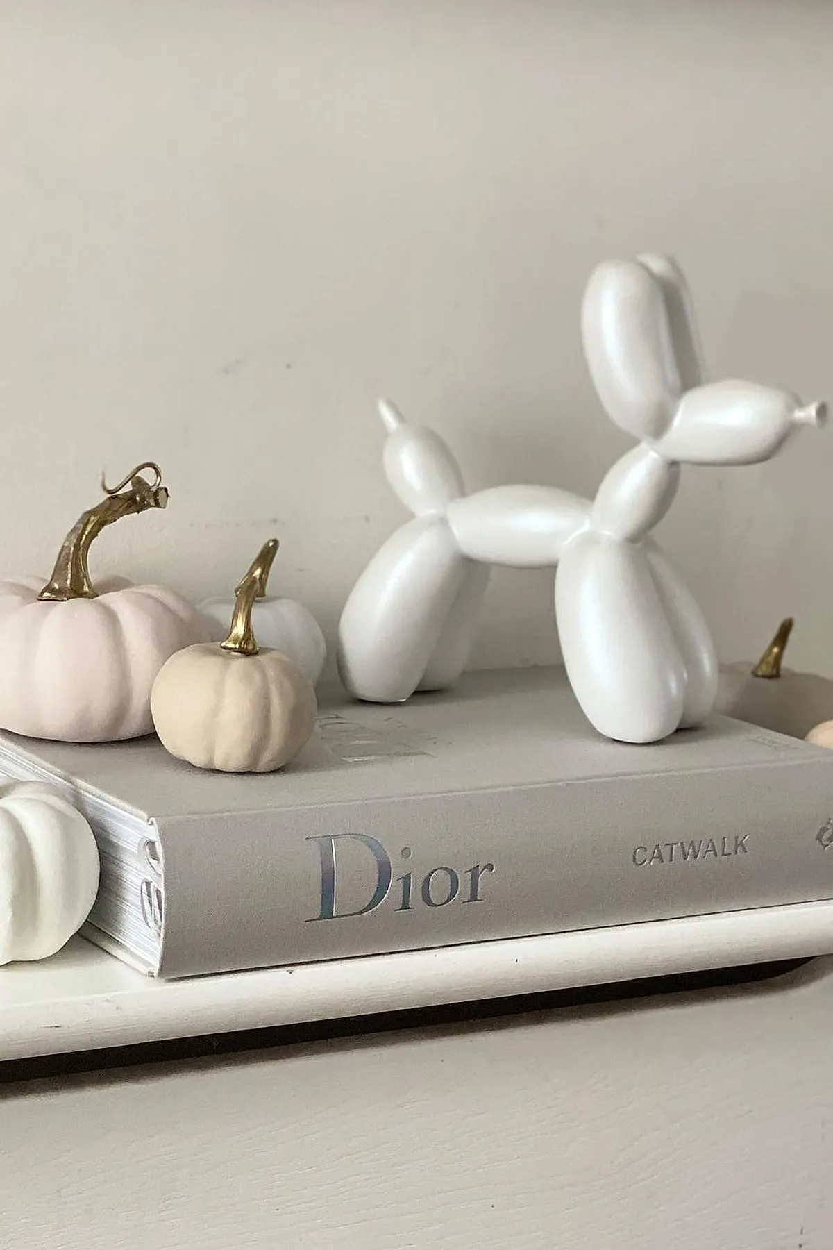 White balloon dog sculpture standing on a grey Dior Catwalk book surrounded by small pale and white pumpkins with shiny gold stems on a light surface against a plain wall.