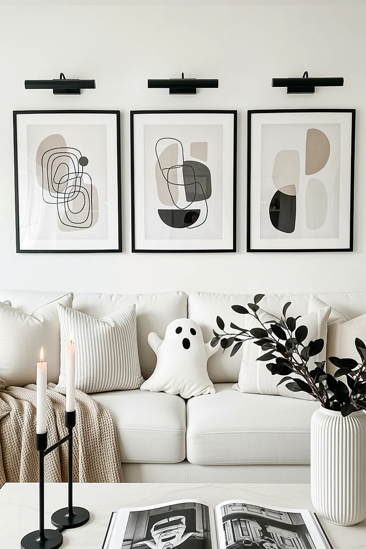 White sofa with beige and striped pillows including a small ghost-shaped pillow, beneath three framed abstract prints in neutral tones, black candleholders with lit white candles on table, and a white ribbed vase holding dark leafy branches.