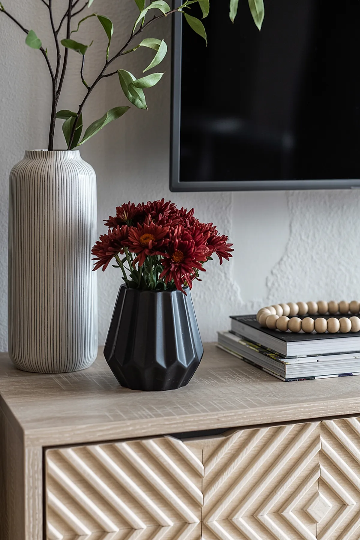 Dark red chrysanthemum flowers arranged in a matte black geometric vase next to a tall white vase with thin vertical stripes, sitting on a carved light wood cabinet with stacked books and wooden bead decor.