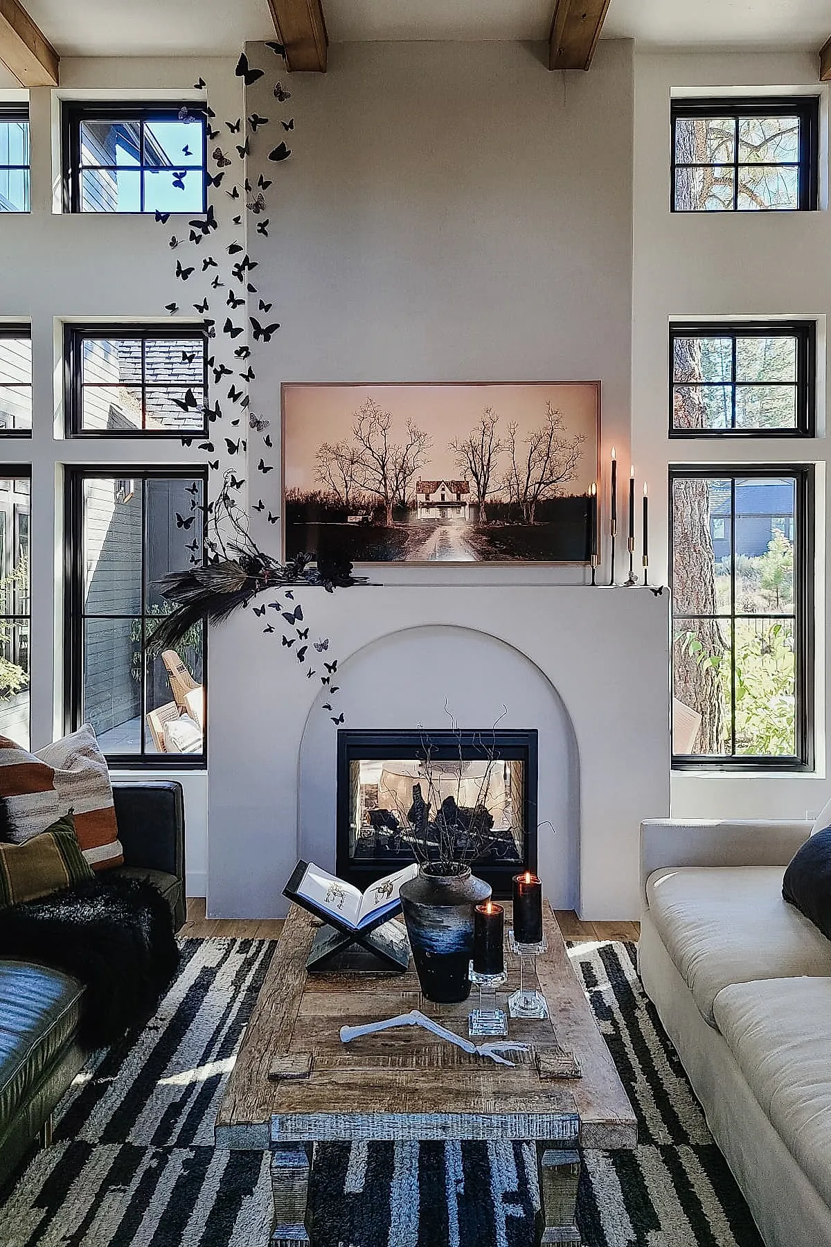 Living room with white fireplace featuring black butterfly wall art climbing vertically beside it, black taper candles on mantle, rustic wooden coffee table with vase holding dried branches, two black candles on glass holders, open book, striped rug, and large windows letting in natural light.