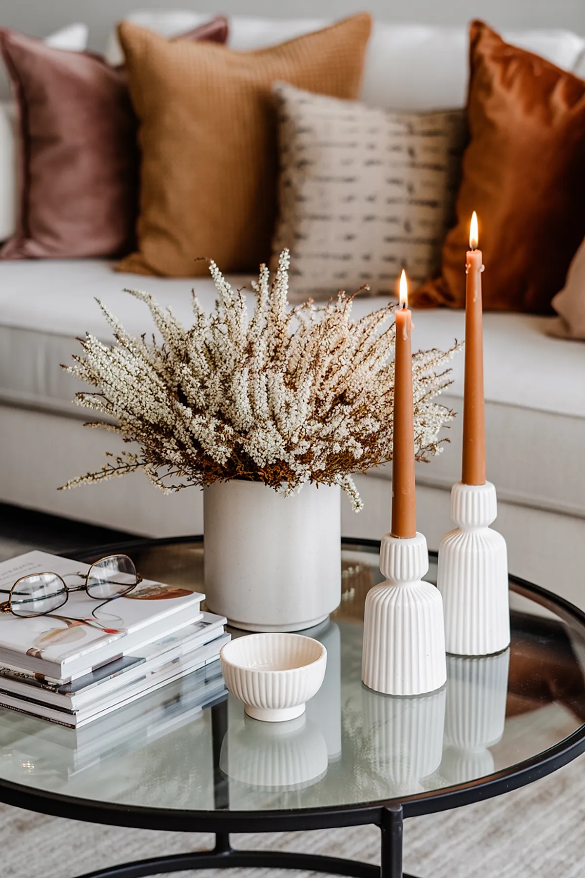 Glass coffee table holding a white ceramic vase with dried delicate white flowers, two lit tan taper candles in ribbed white candlesticks, stack of books with glasses on top, in front of a white sofa with rust, camel, mauve, and patterned beige pillows.