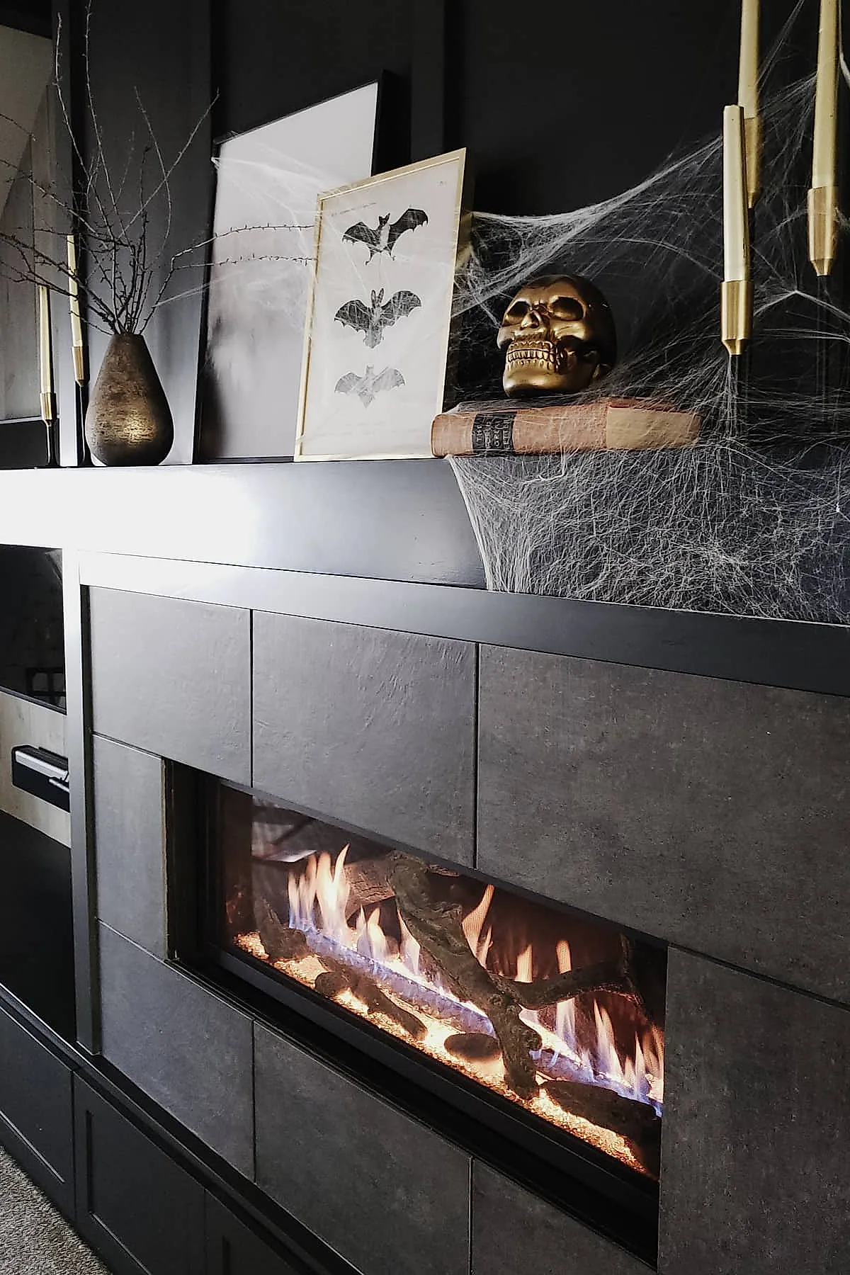 Black modern fireplace mantel decorated with a gold skull atop old books, framed bat art, tall gold taper candles, bare branches in a bronze vase, all accented with white faux spider webs.