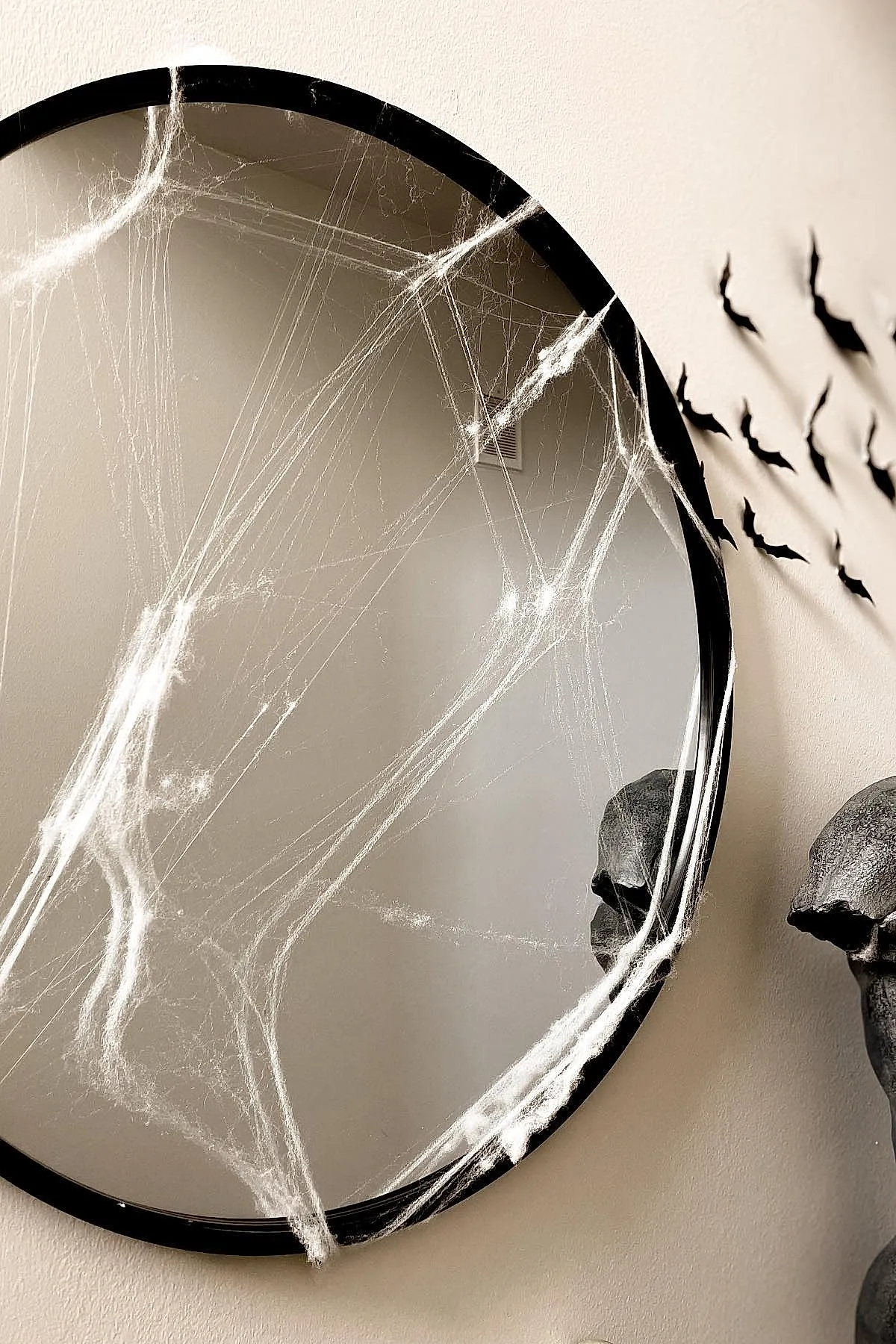 Round black-framed mirror covered in white fake spider webs next to a beige wall decorated with black bat silhouettes and part of a dark skull sculpture.