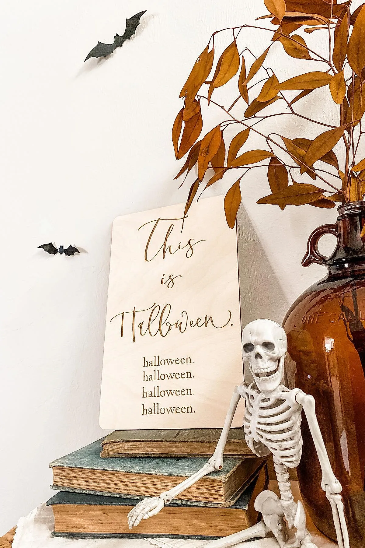 Stack of worn hardcover books topped with a cream-colored sign reading 'This is Halloween', next to a small white plastic skeleton and an amber glass vase holding brown dried leaves, against a white wall decorated with two black bat cutouts.