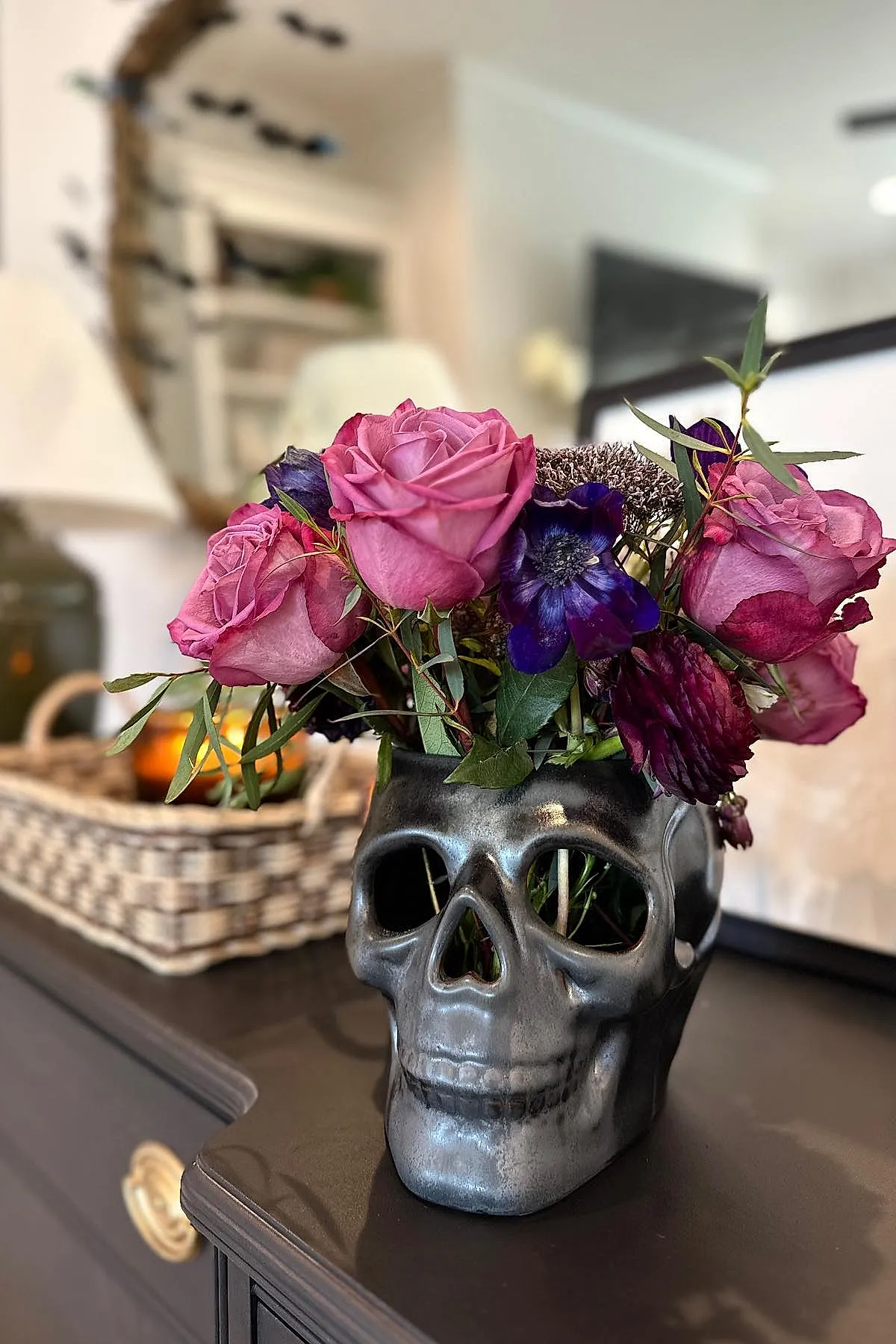 Metallic silver skull-shaped vase filled with vibrant purple and pink roses and other flowers placed on a dark wooden surface.