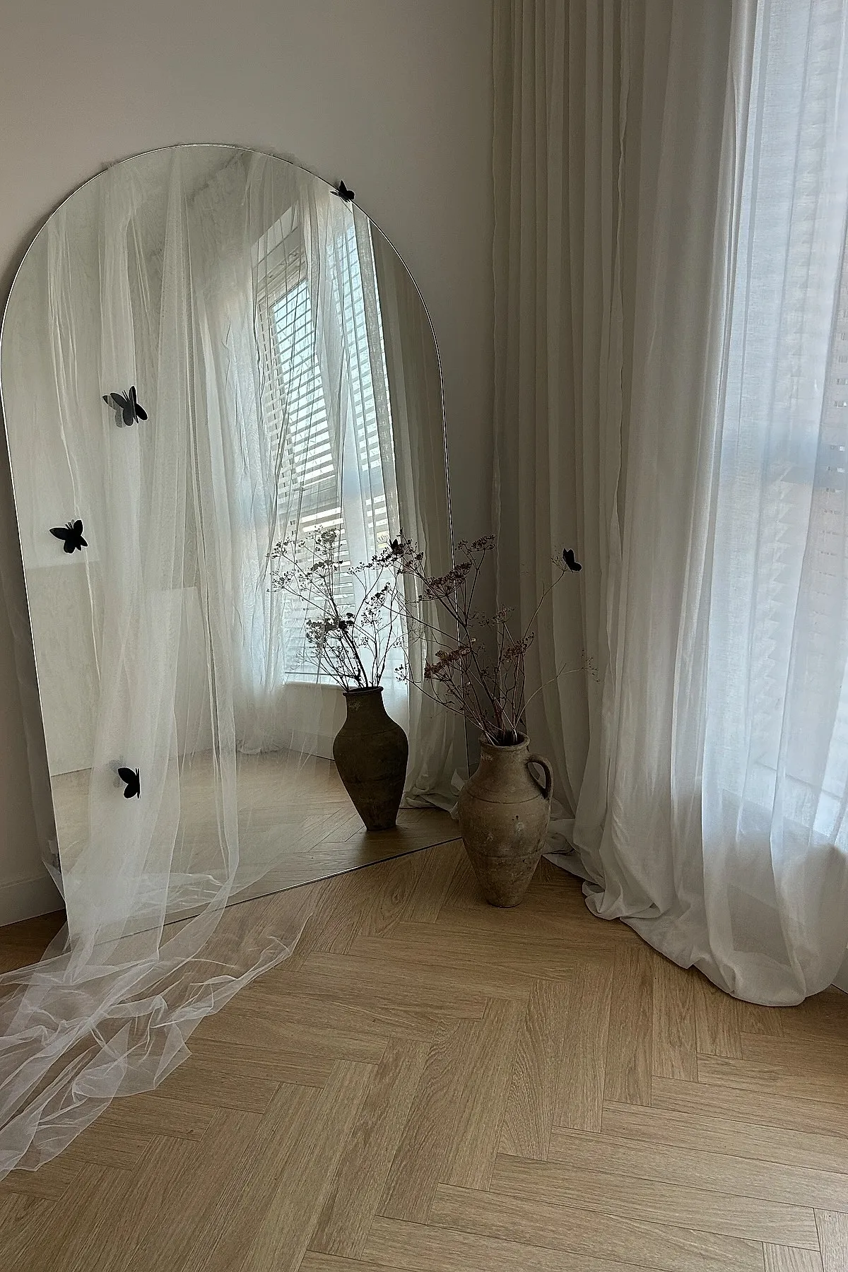 Large arched mirror leaning against wall with sheer white fabric draped over it and small black butterfly decorations attached. Beside the mirror is a rustic clay vase holding dried branches, all set on light wood herringbone floor next to white sheer curtains.
