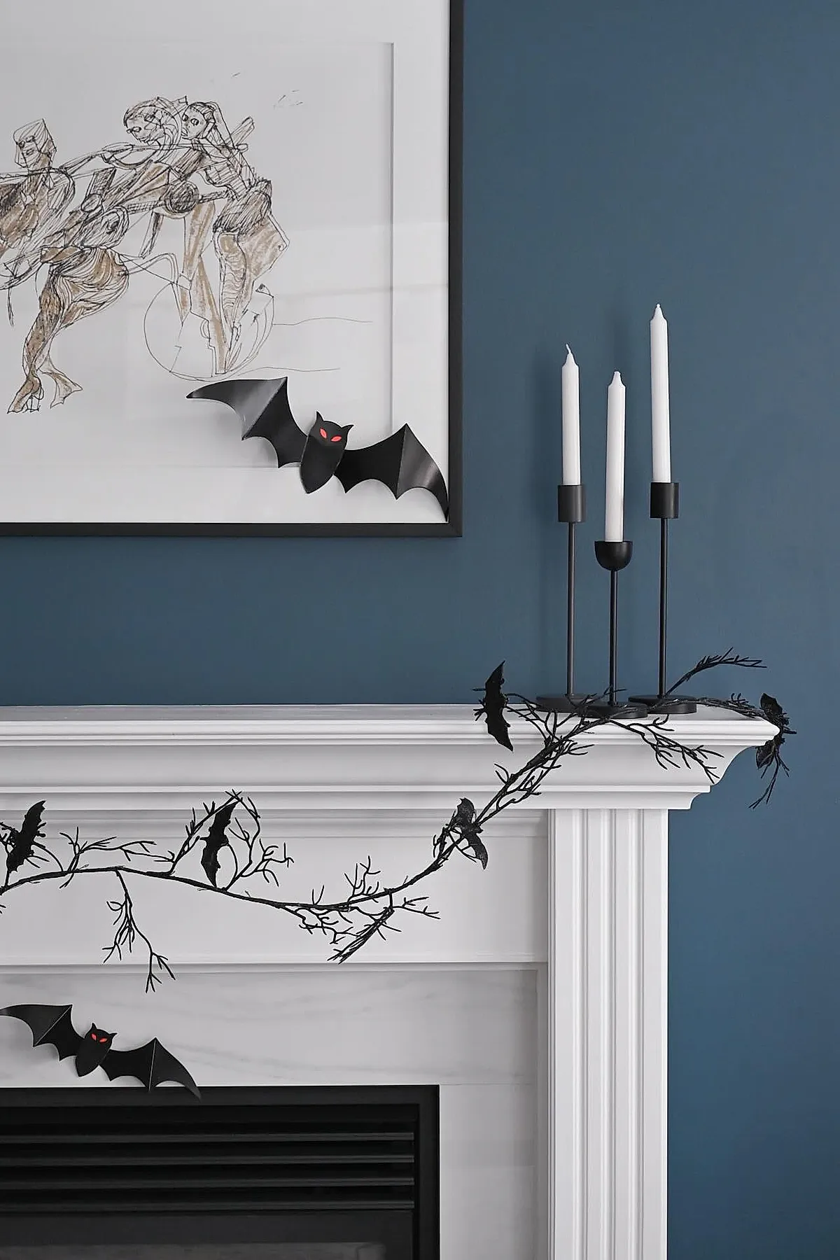 White fireplace mantle against blue wall decorated with black metal candle holders holding three white taper candles, black bat garland with branch-like details draped across the mantle, and a framed abstract artwork featuring small black bats with red eyes attached to its bottom corner.