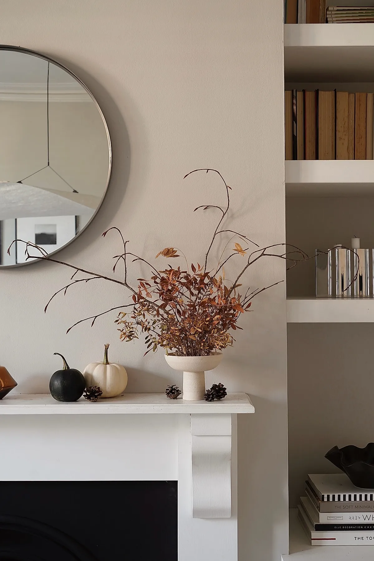 White pedestal vase holding dried brown branches and leaves on white mantel alongside small black and white pumpkins and scattered pinecones against a beige wall with round mirror above.