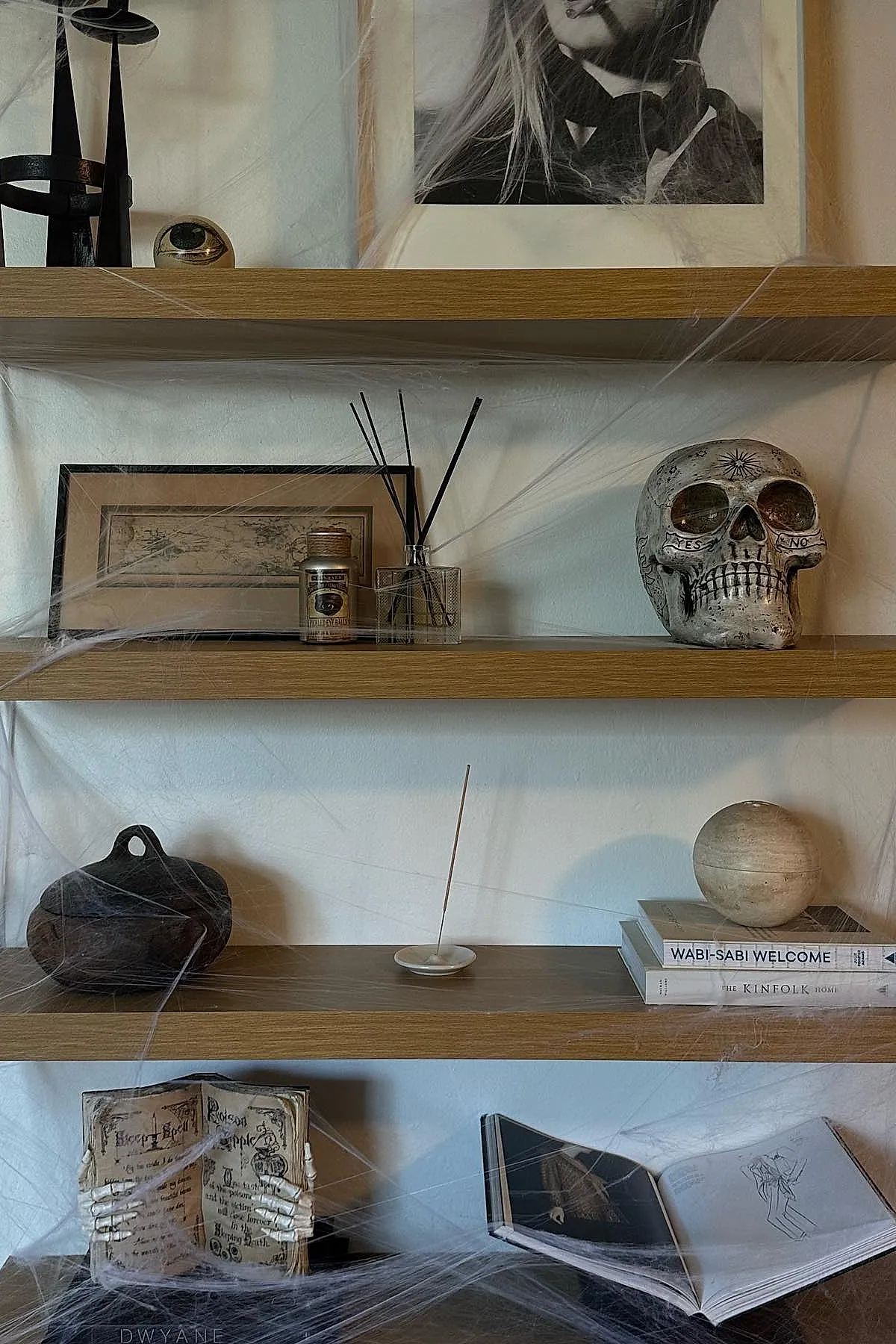 Wooden floating shelves decorated with black candle holders, framed art, skull sculpture, diffuser sticks in glass jar, pottery piece, incense stick on dish, stack of books topped by round stone ball, skeleton hand holding open book prop, open sketchbook, all draped with thin white fake cobwebs.