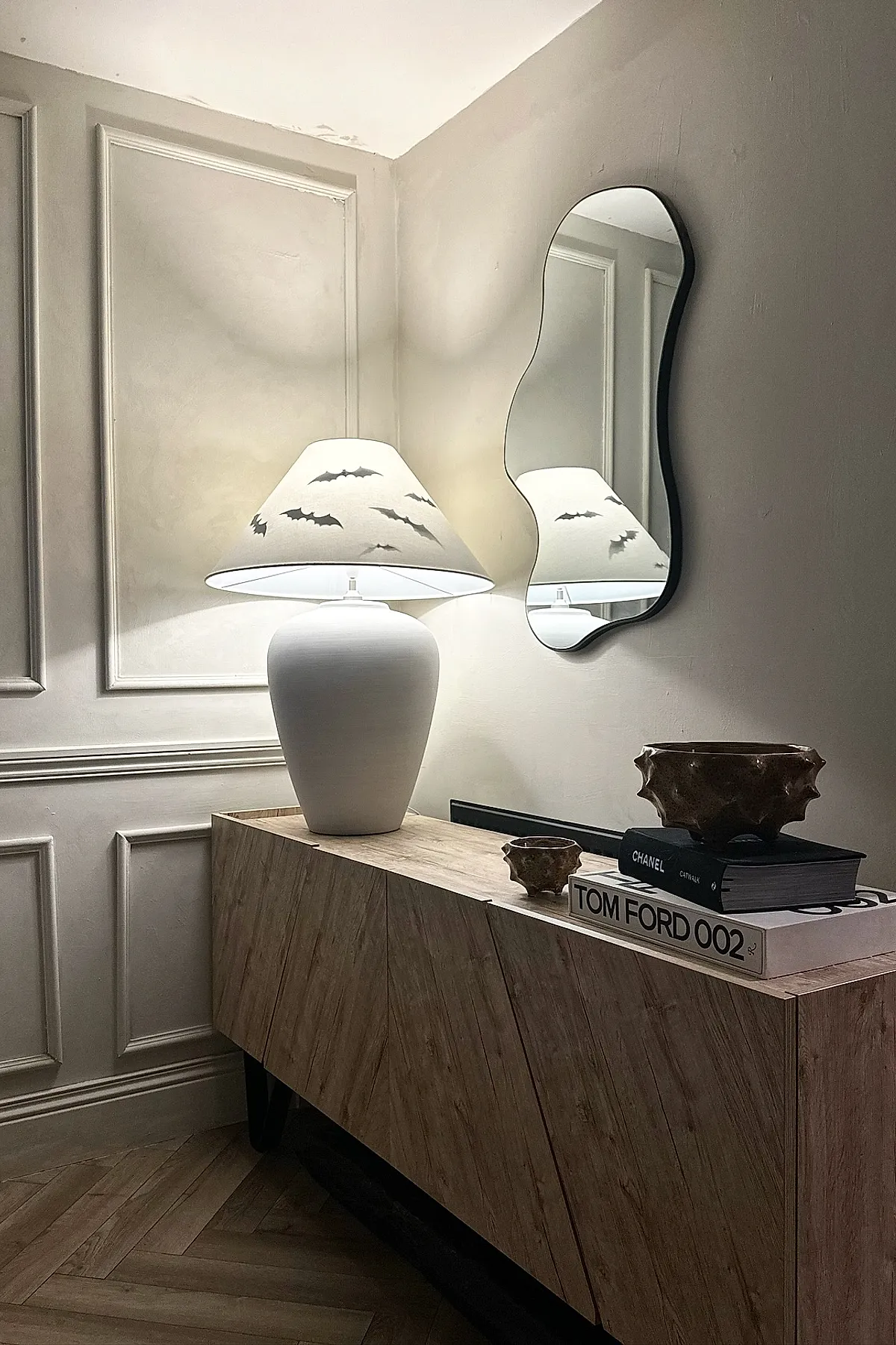 White table lamp with bat silhouette patterned shade on wooden console next to wavy black-framed mirror mounted on beige wall, accompanied by two brown textured ceramic bowls and stacked fashion books.