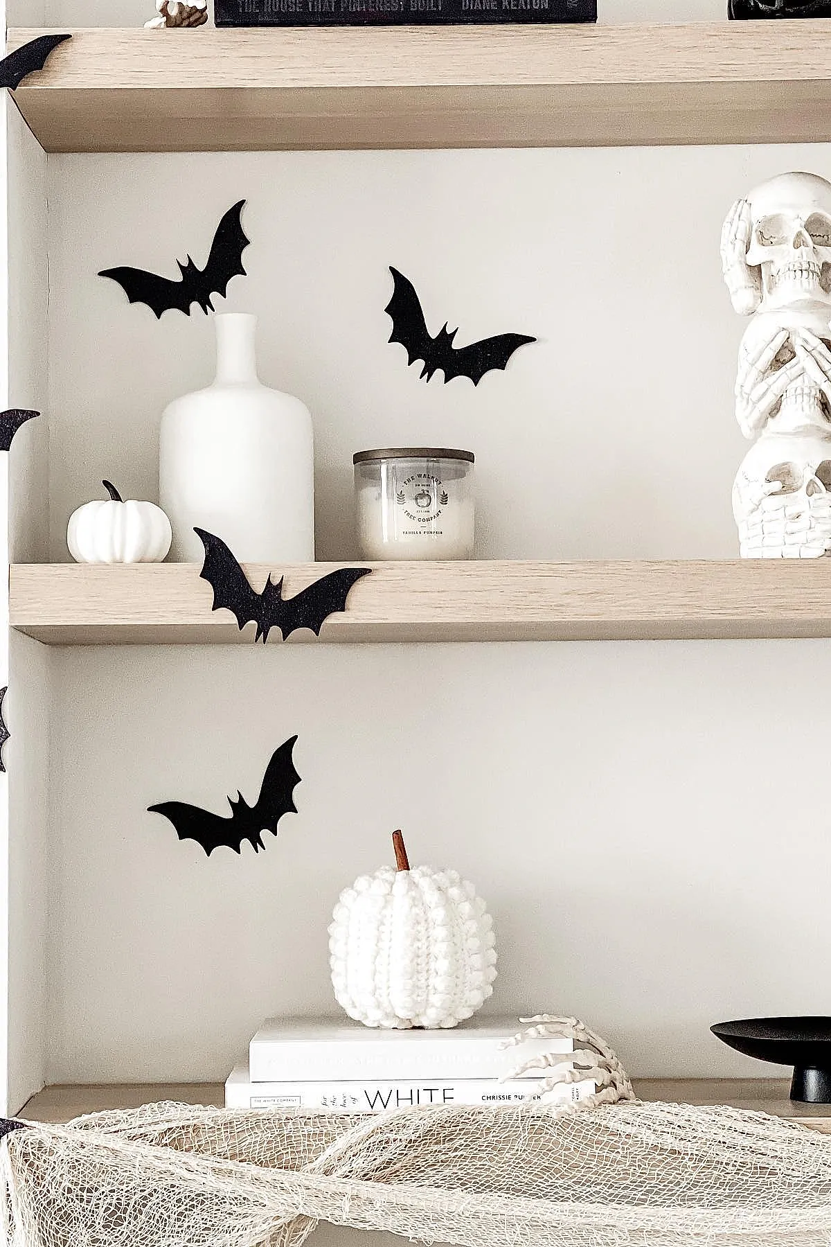 Light wood shelves decorated with black bat wall decals, small white pumpkins, a white ceramic bottle, candle in glass jar, and stacked white skull sculptures on a neutral background.