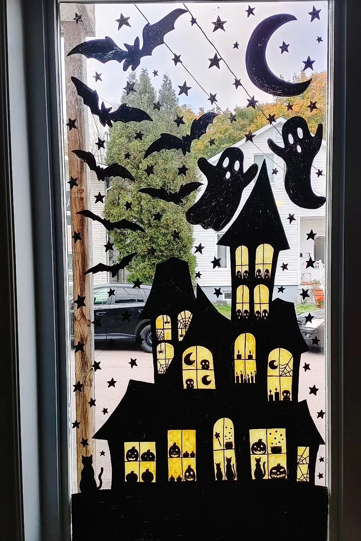 Black silhouette of a haunted house with glowing yellow windows, surrounded by flying bats, ghosts, stars, and a crescent moon on a clear window.