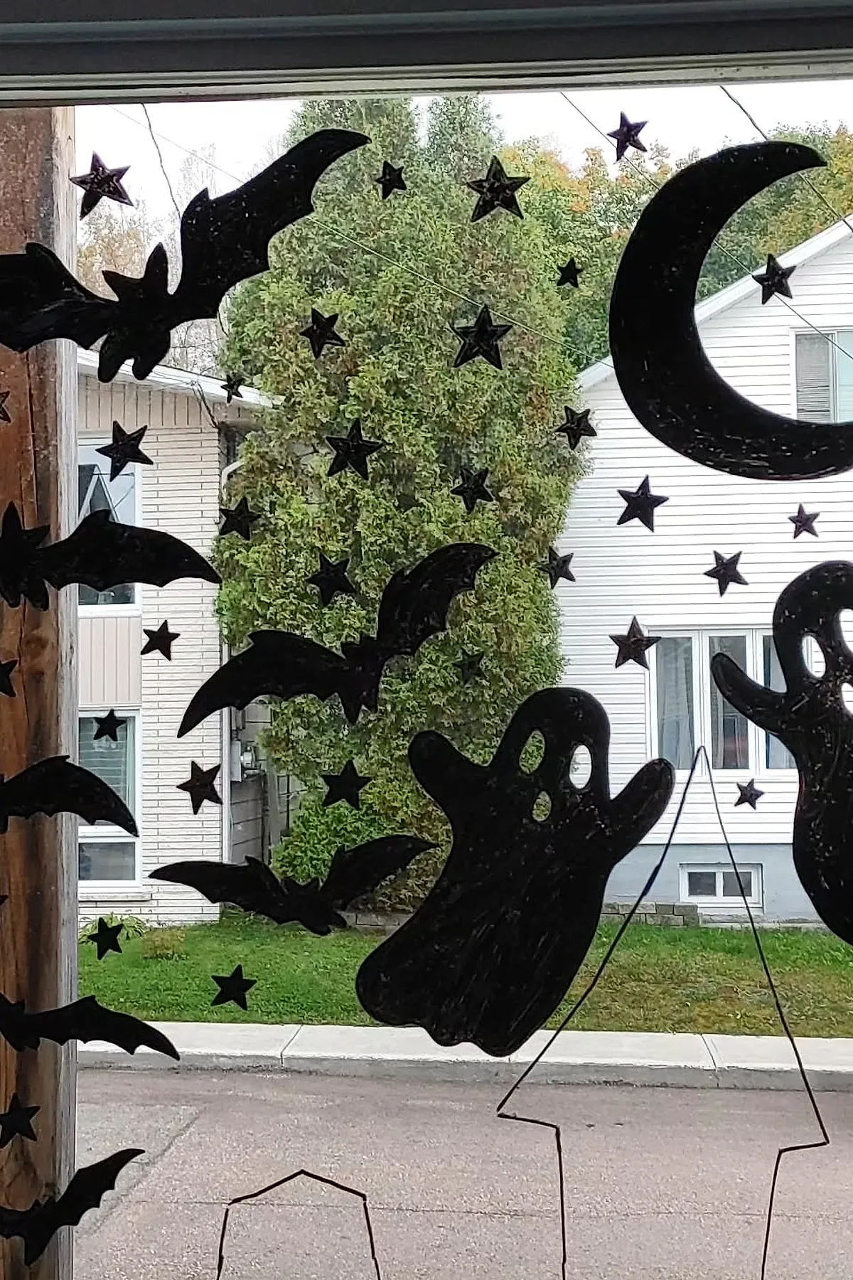 Black paper cutouts of flying bats, two ghost figures, numerous small stars, and a large crescent moon arranged on a clear window with outdoor view.