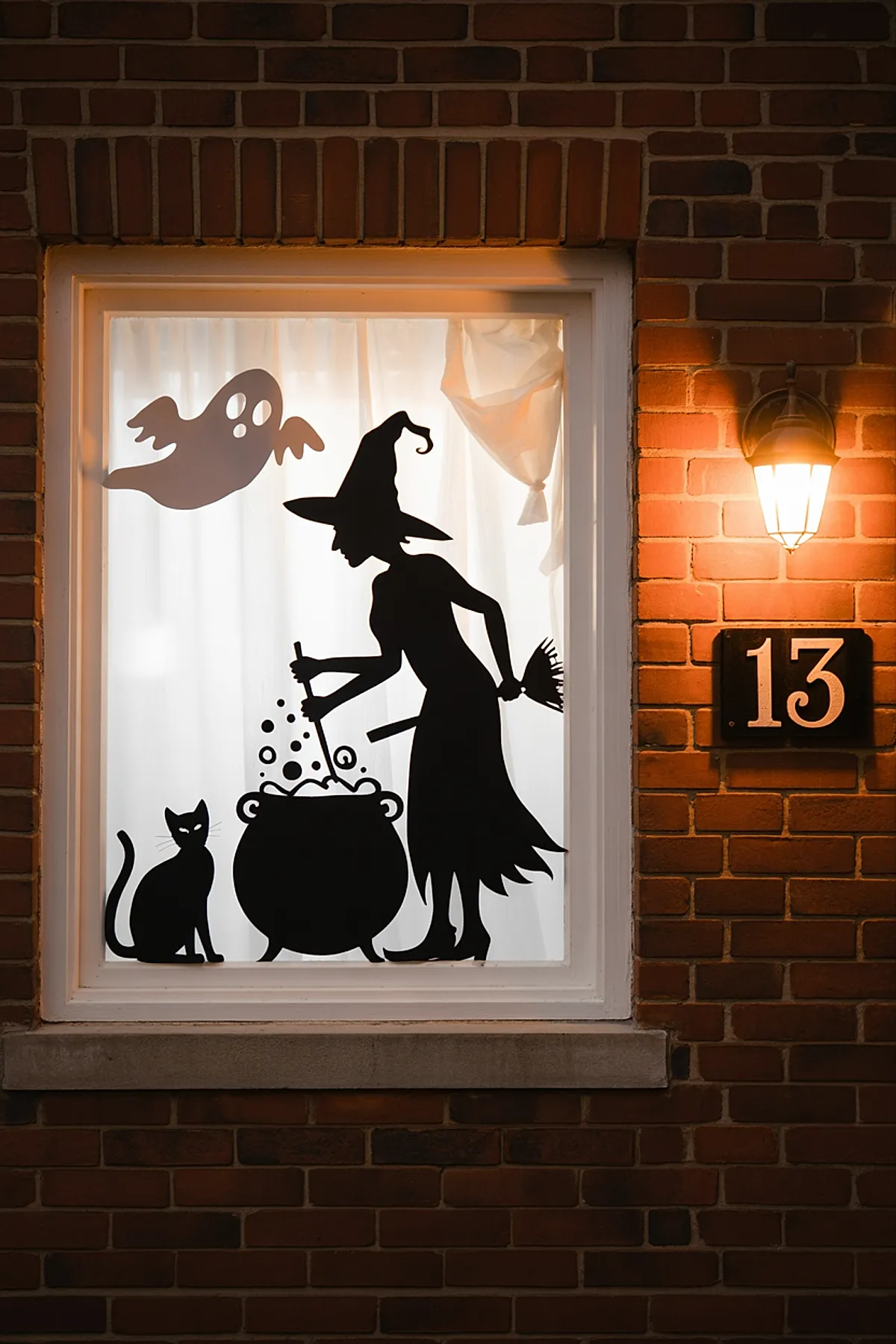 Window with white curtains showing black silhouettes of a witch stirring a bubbling cauldron, a sitting cat, and a ghost floating above on brick exterior wall with lit porch light and house number 13.