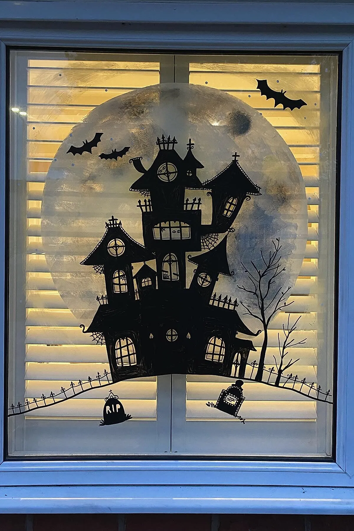Window display featuring a black silhouette of a crooked haunted house on a hill with bare trees, gravestones, and flying bats against a painted gray full moon background.