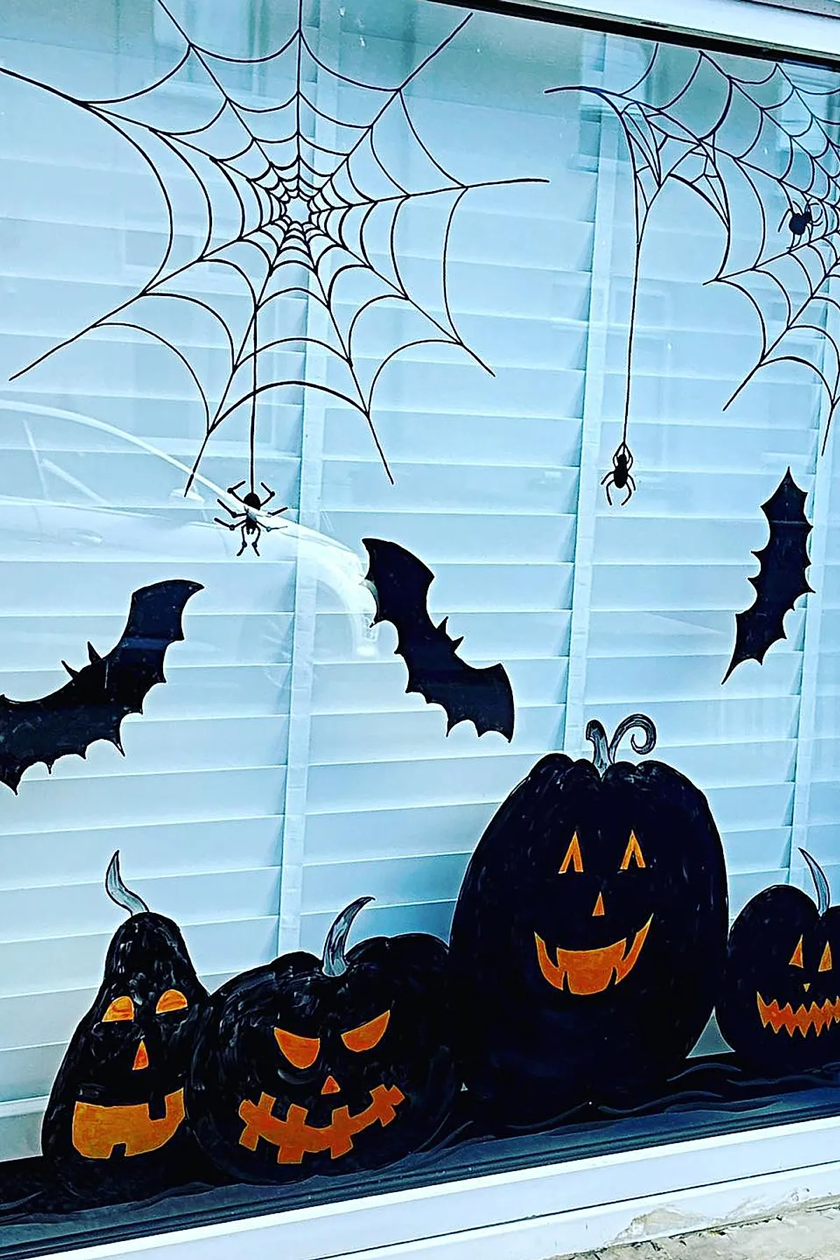 Window decorated with black painted pumpkins showing orange jack-o-lantern faces, black bats flying, large spider webs, and small spiders hanging from web strands.