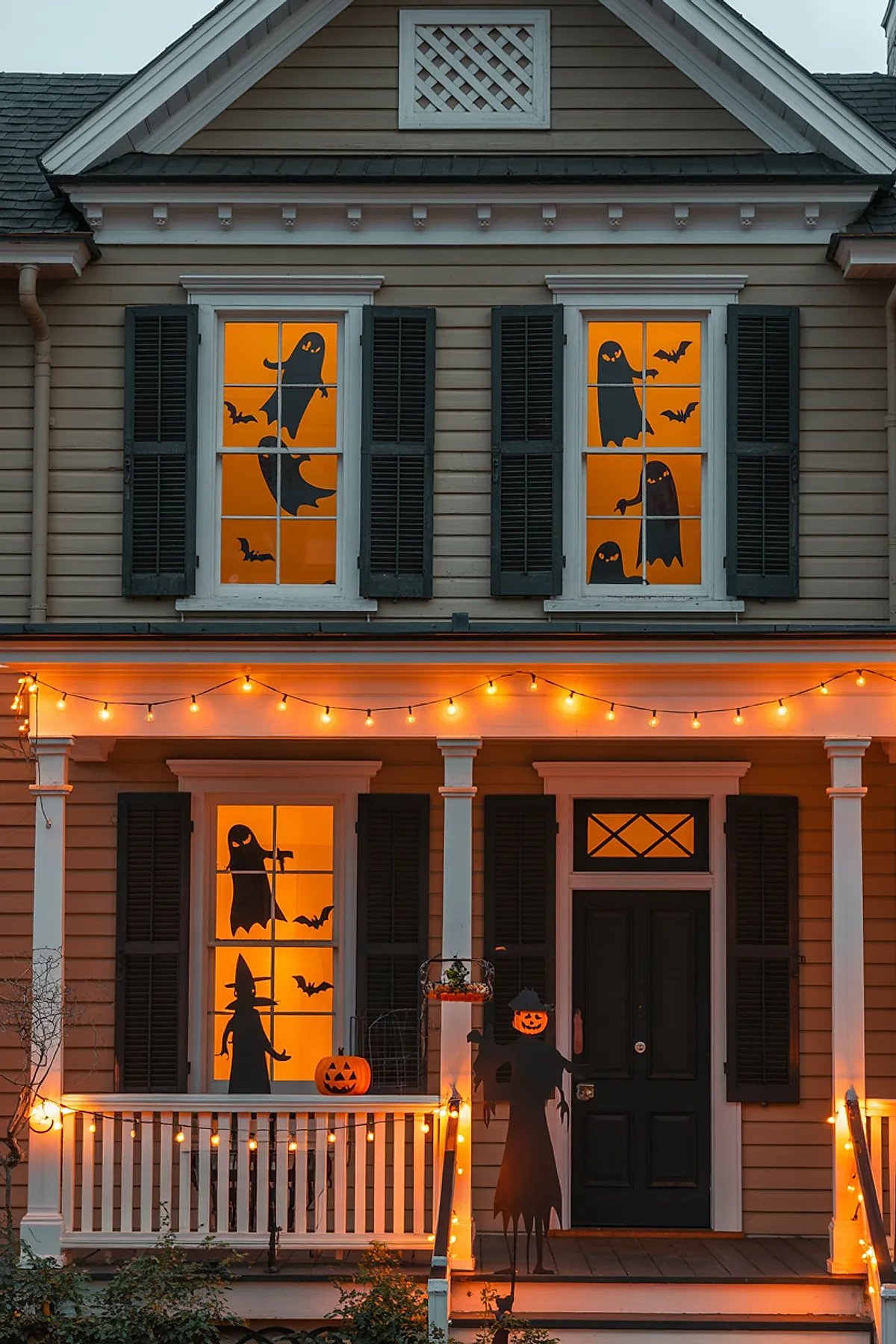 House with four windows featuring black silhouette cutouts of ghosts, bats, and witches illuminated by orange light from inside, decorated with string lights on porch.