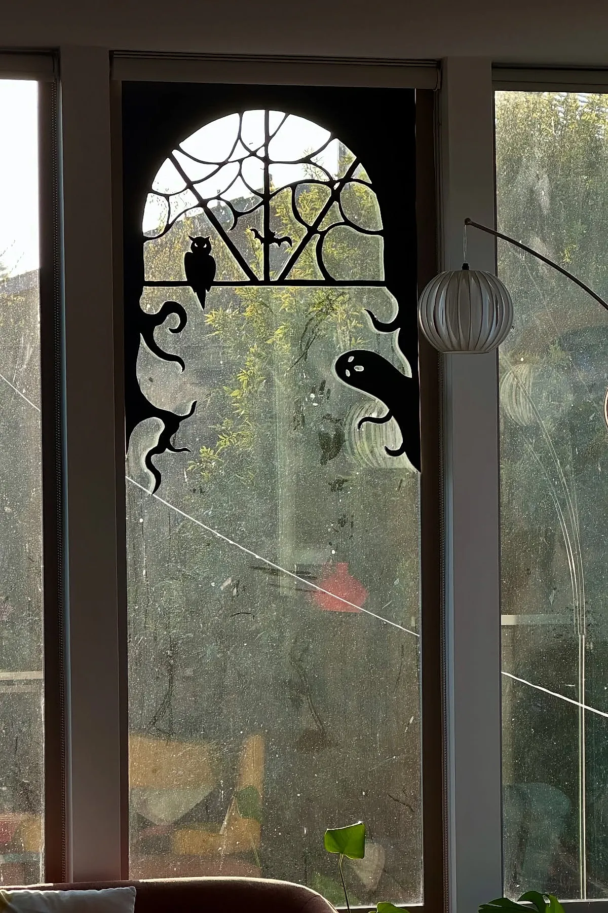 Black Halloween window cutout featuring an owl on a branch, a spider web with a small bat, twisted tree branches, and a ghost against a tall arched glass window.