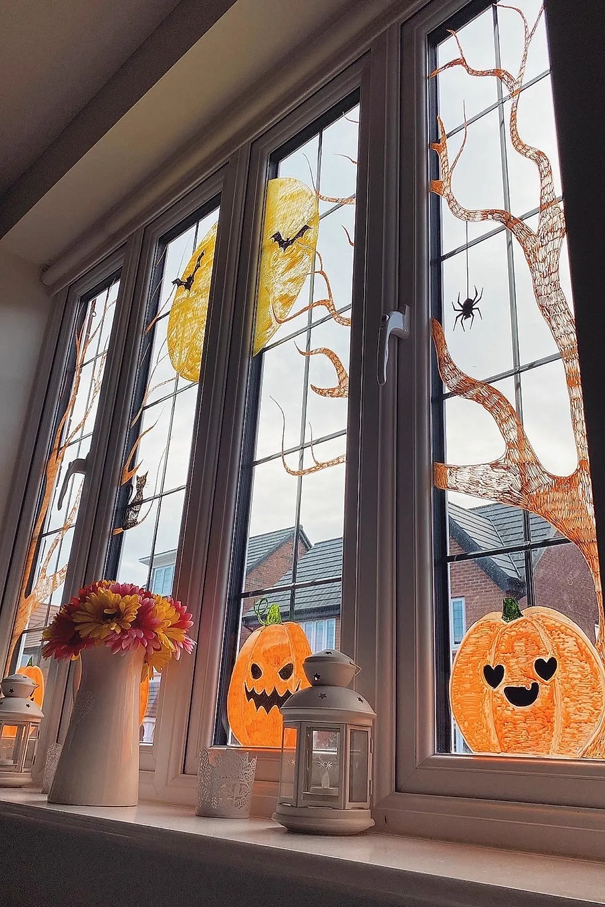 Four-pane window decorated with orange hand-drawn pumpkins with different faces, leafless brown trees, yellow moons, black bats, and hanging spiders on clear glass.