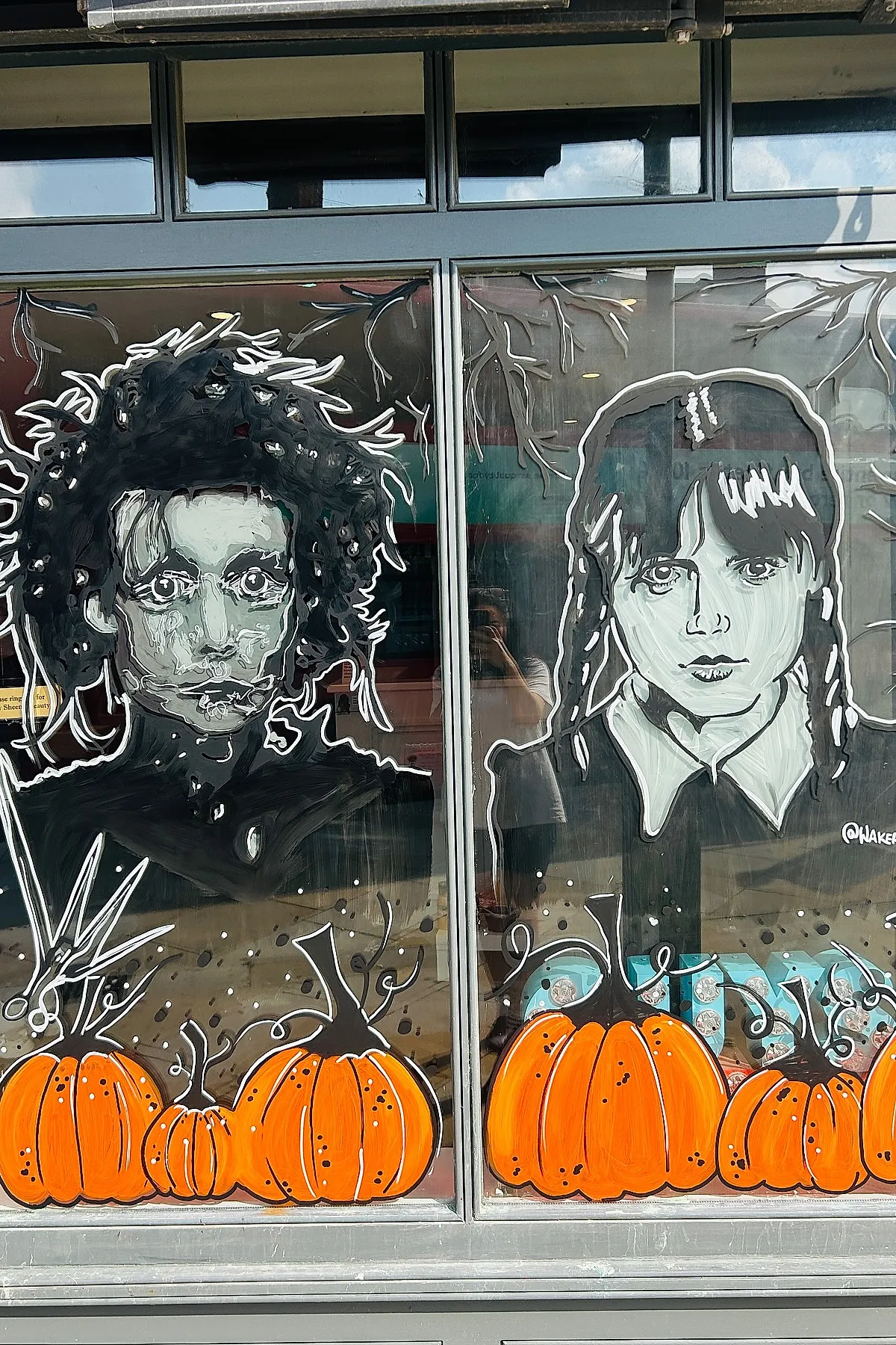 Black and white painted portraits of Edward Scissorhands and Wednesday Addams on a window, surrounded by bright orange pumpkins along the bottom edge.
