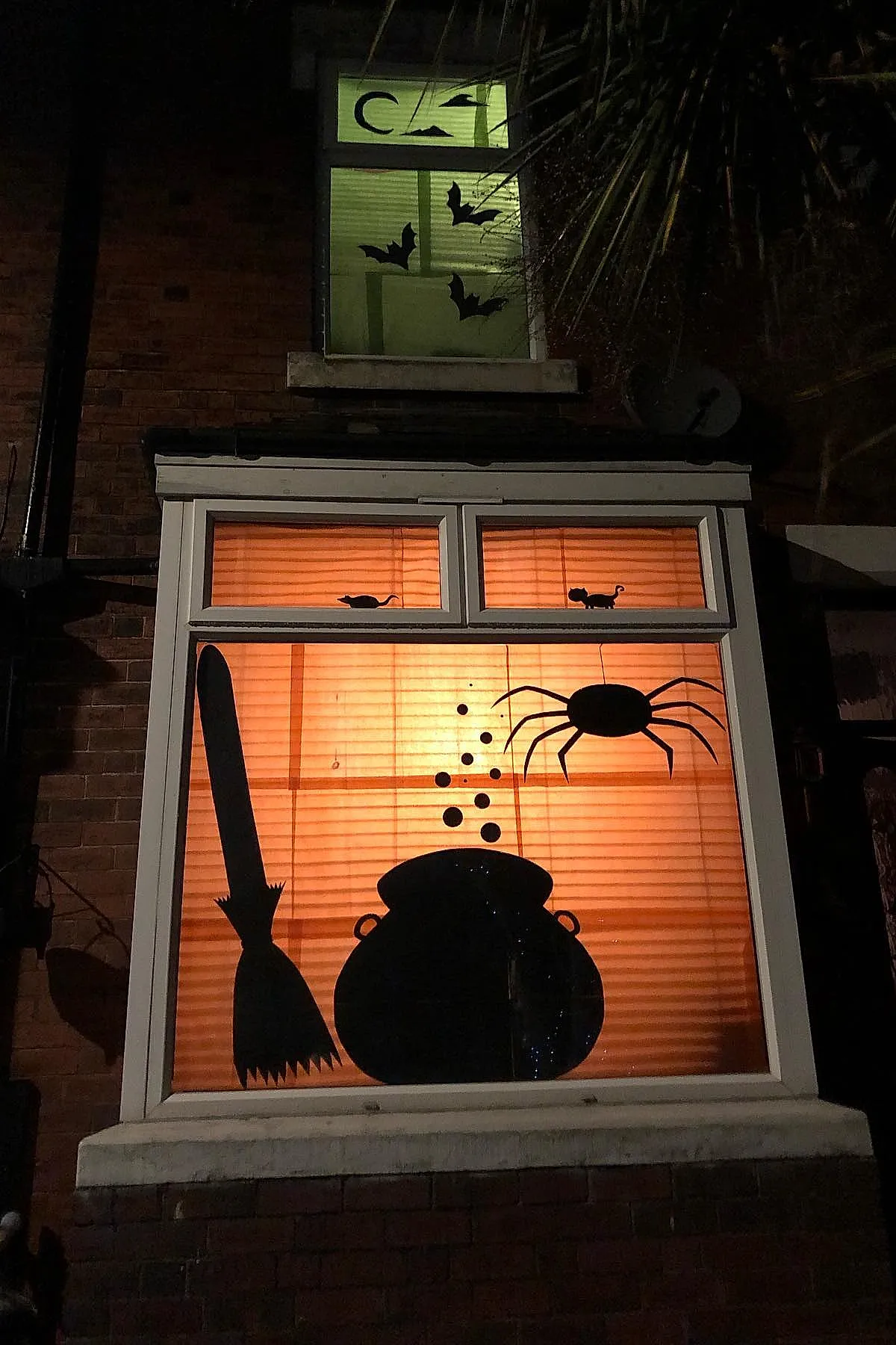 Orange backlit window with black silhouette cutouts including a broom, bubbling cauldron with bubbles rising, a large spider hanging down, and small mouse shapes in upper smaller window panes.