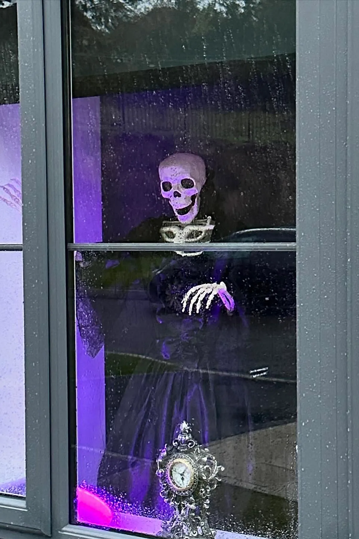 Skeleton figure wearing black clothes displayed behind a rain-speckled window lit with purple light, with an ornate vintage clock placed at the bottom.