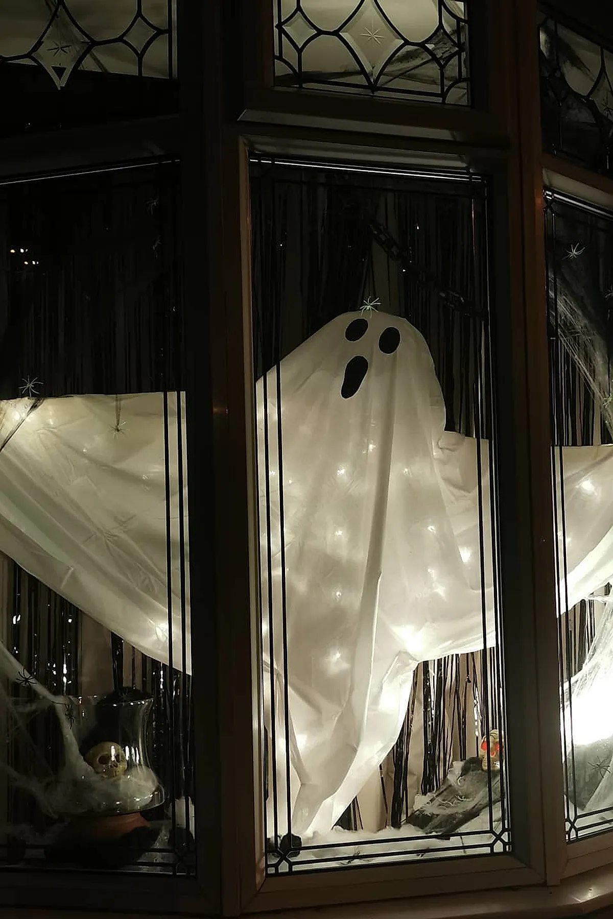 Window display featuring a glowing white ghost made from fabric lit with warm white string lights, surrounded by faux cobwebs and small skull decorations behind glass panes.