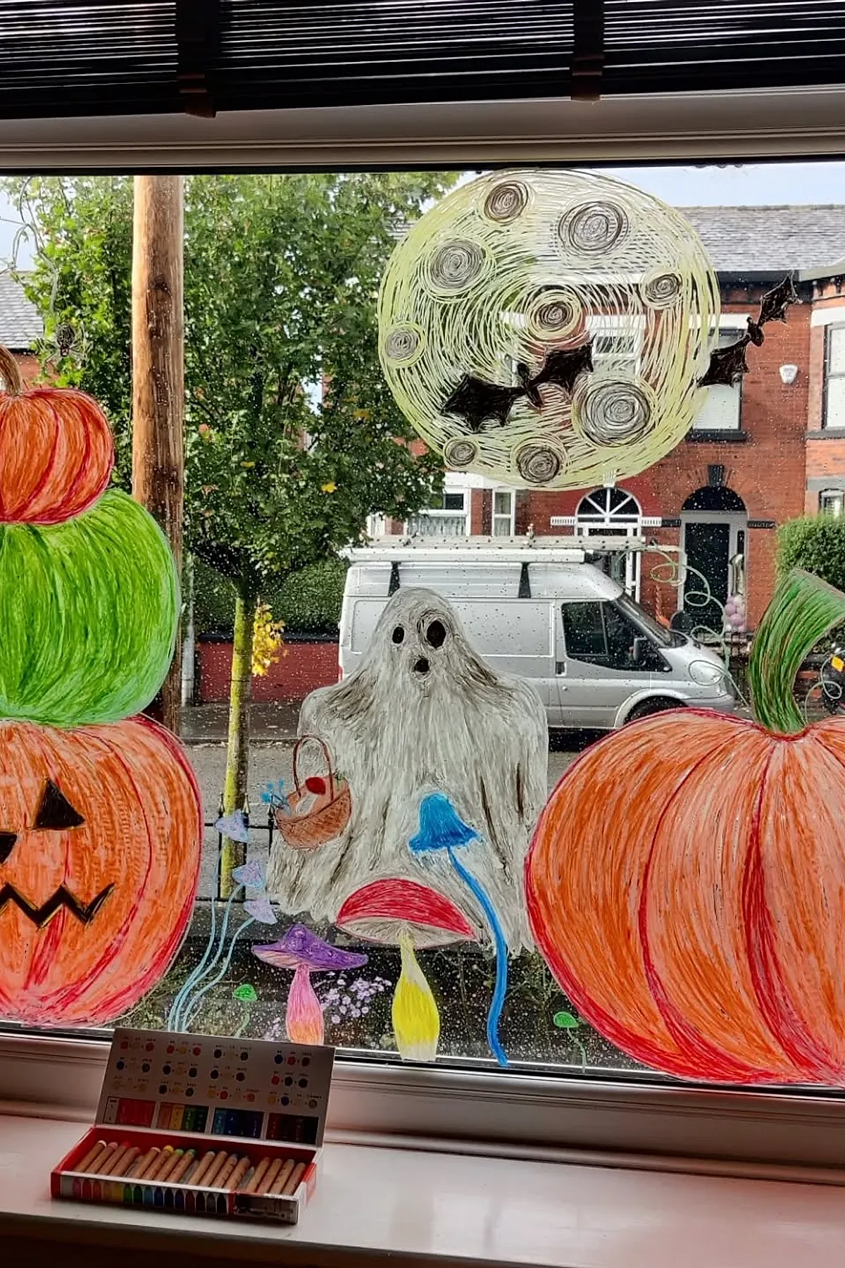 Colorful Halloween window painting featuring stacked orange and green pumpkins, a white ghost holding a basket, multicolored mushrooms at the bottom, and a swirling yellow full moon with black bats flying across it.