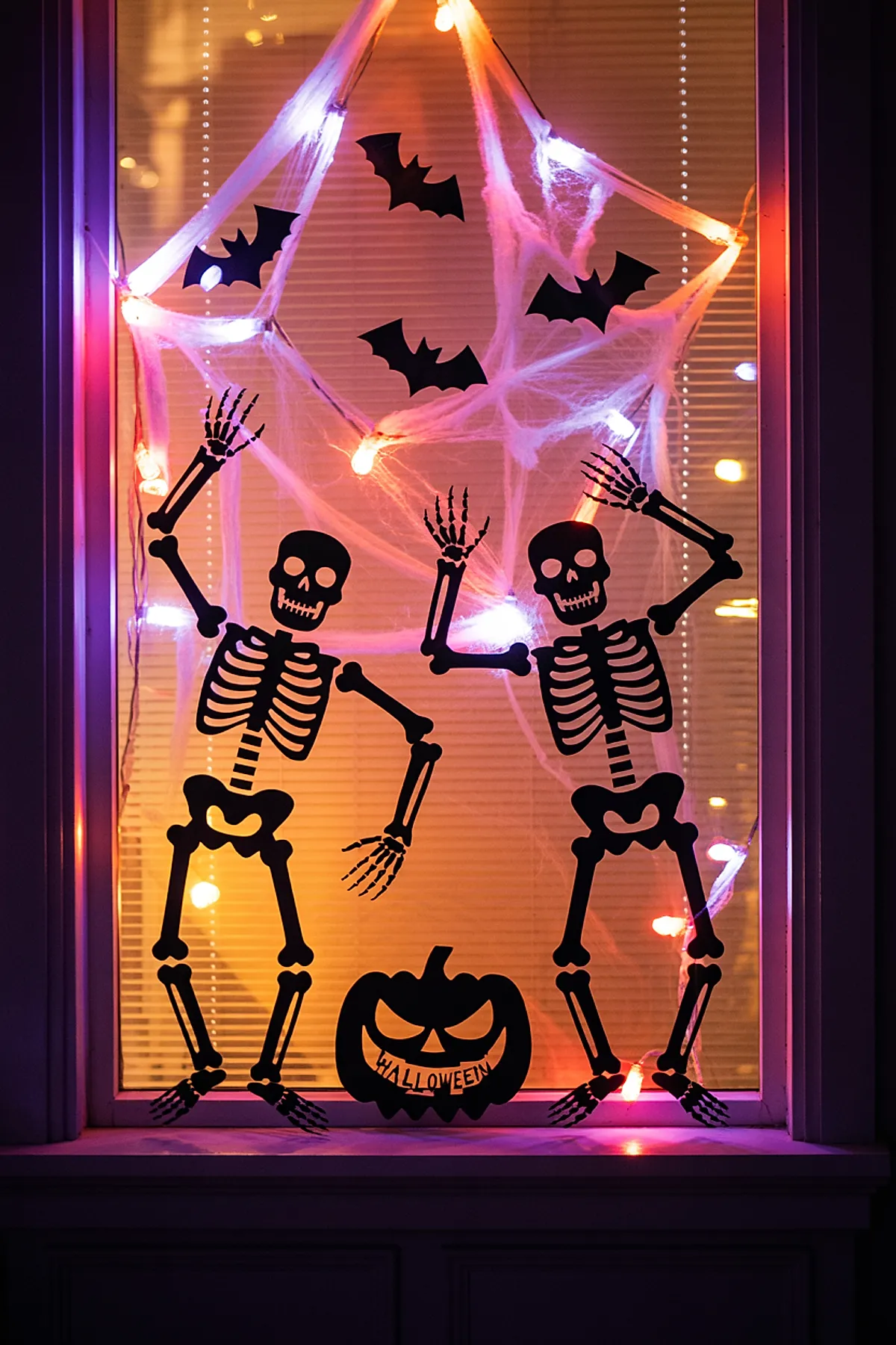 Window decorated with black skeleton cutouts in dancing poses, black bats, and a glowing white spider web made from stretched fabric with multicolored string lights, plus a black pumpkin silhouette with Halloween text at the bottom.