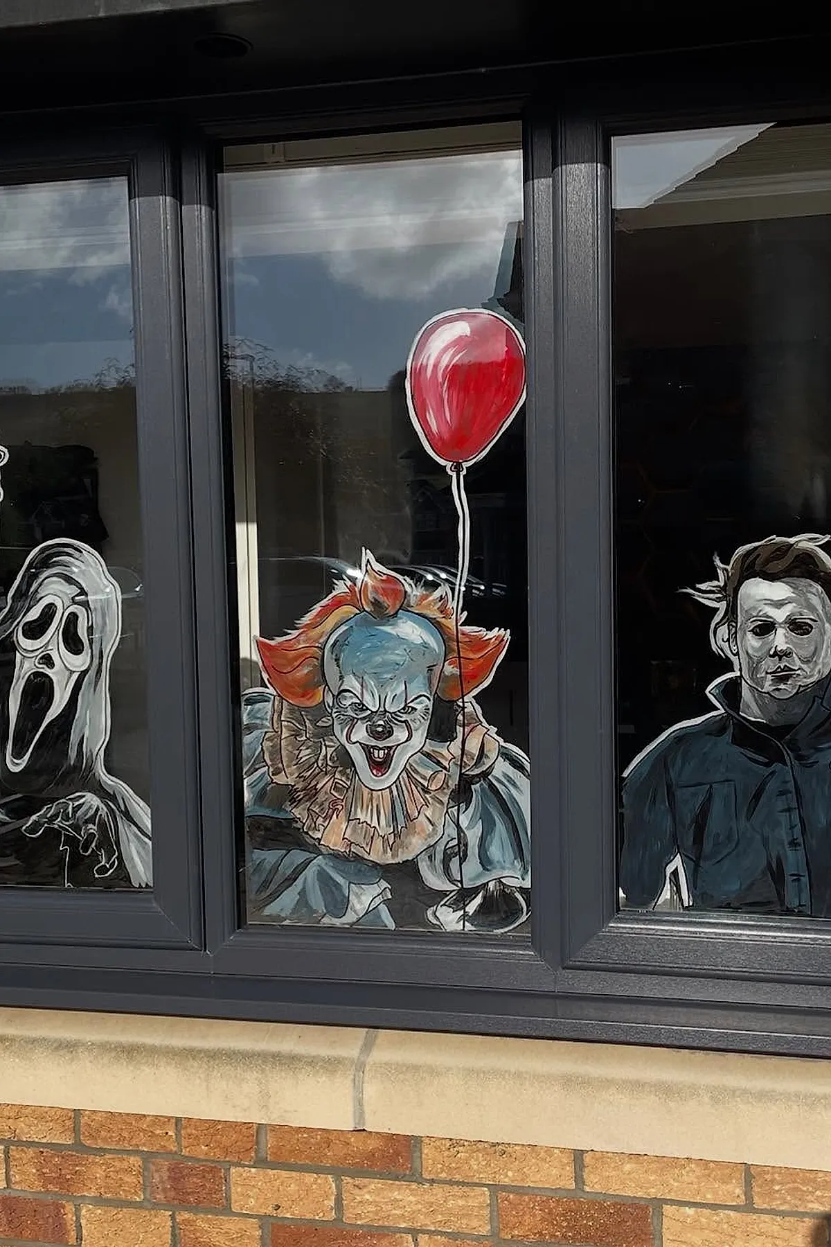 Three painted horror movie characters on black-framed windows including a ghost face mask, a clown with orange hair holding a red balloon, and a masked figure in dark clothing.