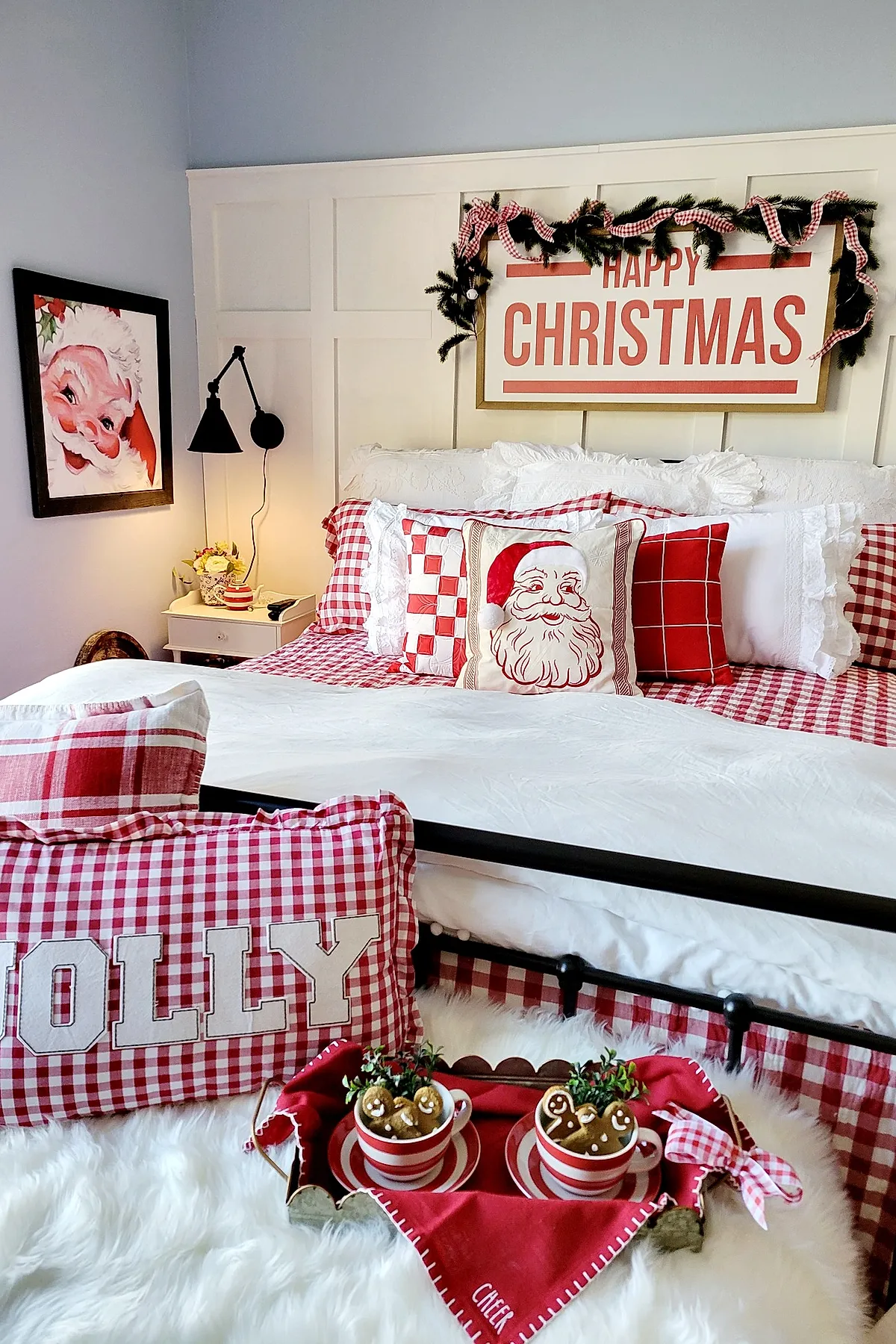 Christmas bedroom decorated with red and white gingham bedding and pillows, a white quilt, a 'Happy Christmas' sign above the headboard adorned with greenery and red gingham ribbon, a framed Santa Claus portrait on the wall next to a black swing-arm lamp, and a cozy tray with striped mugs and gingerbread men on a fluffy white rug at the foot of the bed.