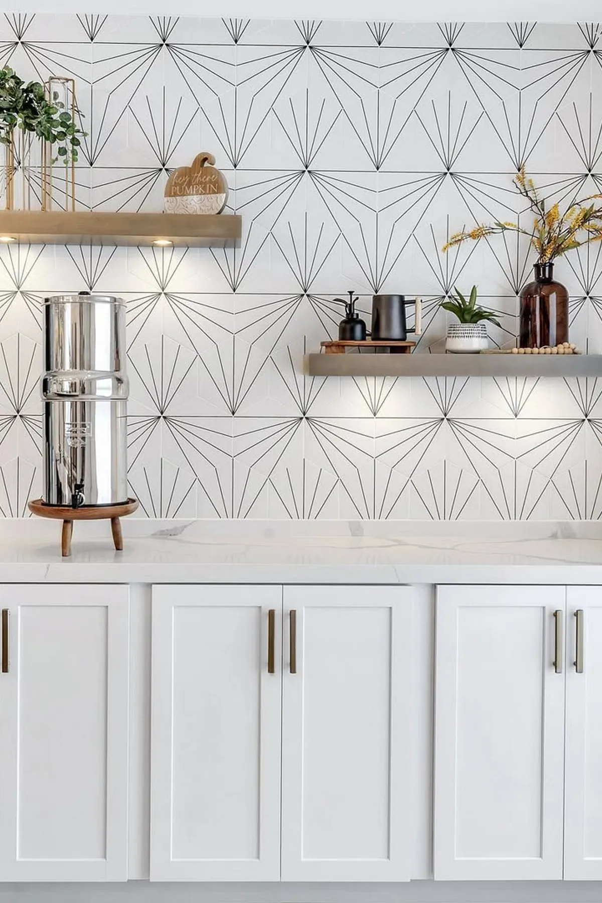 White cabinets with brass handles and a geometric patterned wallpaper behind wooden shelves.