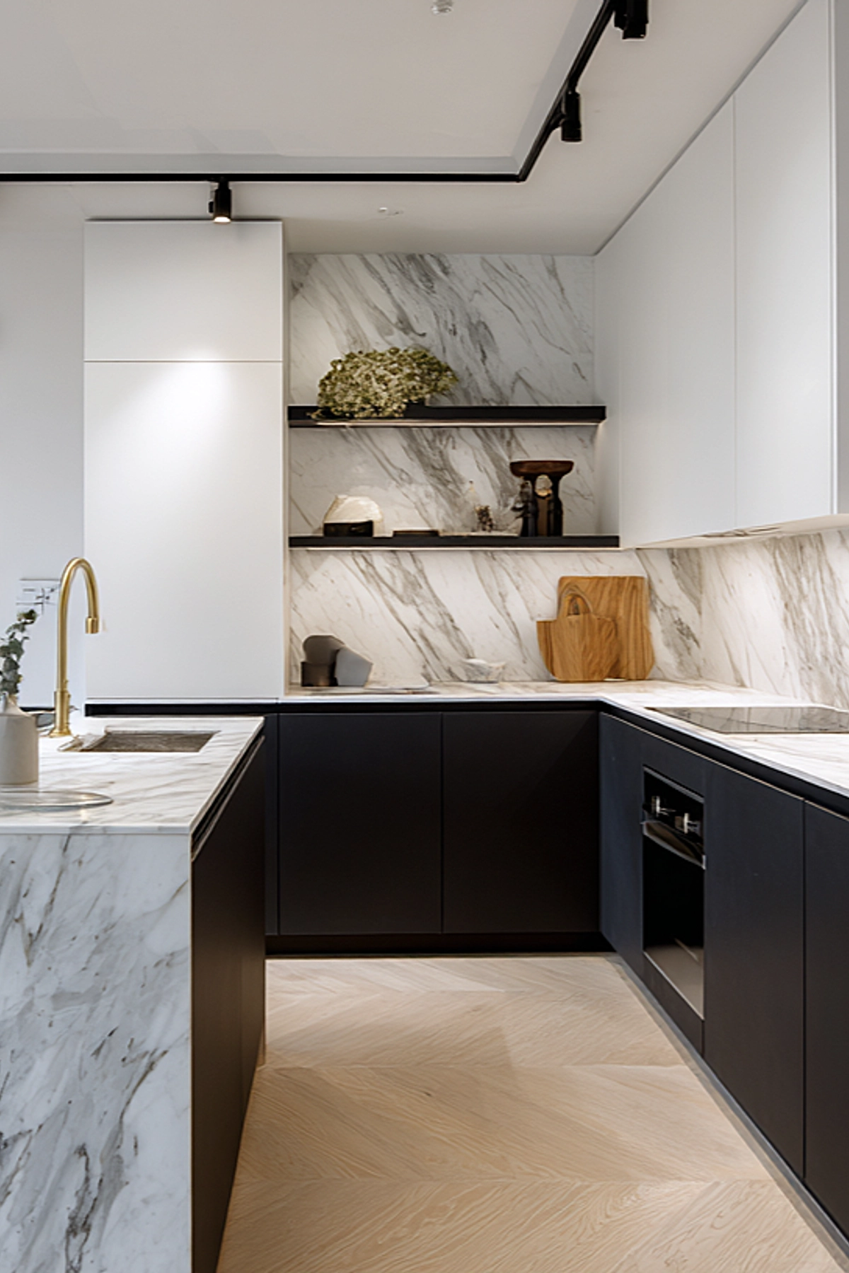 Small modern kitchen with marble countertops and backsplash, dark lower cabinets, and white upper cabinets. Open shelves display decor, while a brass faucet adds warmth. The U-shaped layout maximizes workspace in a compact footprint.