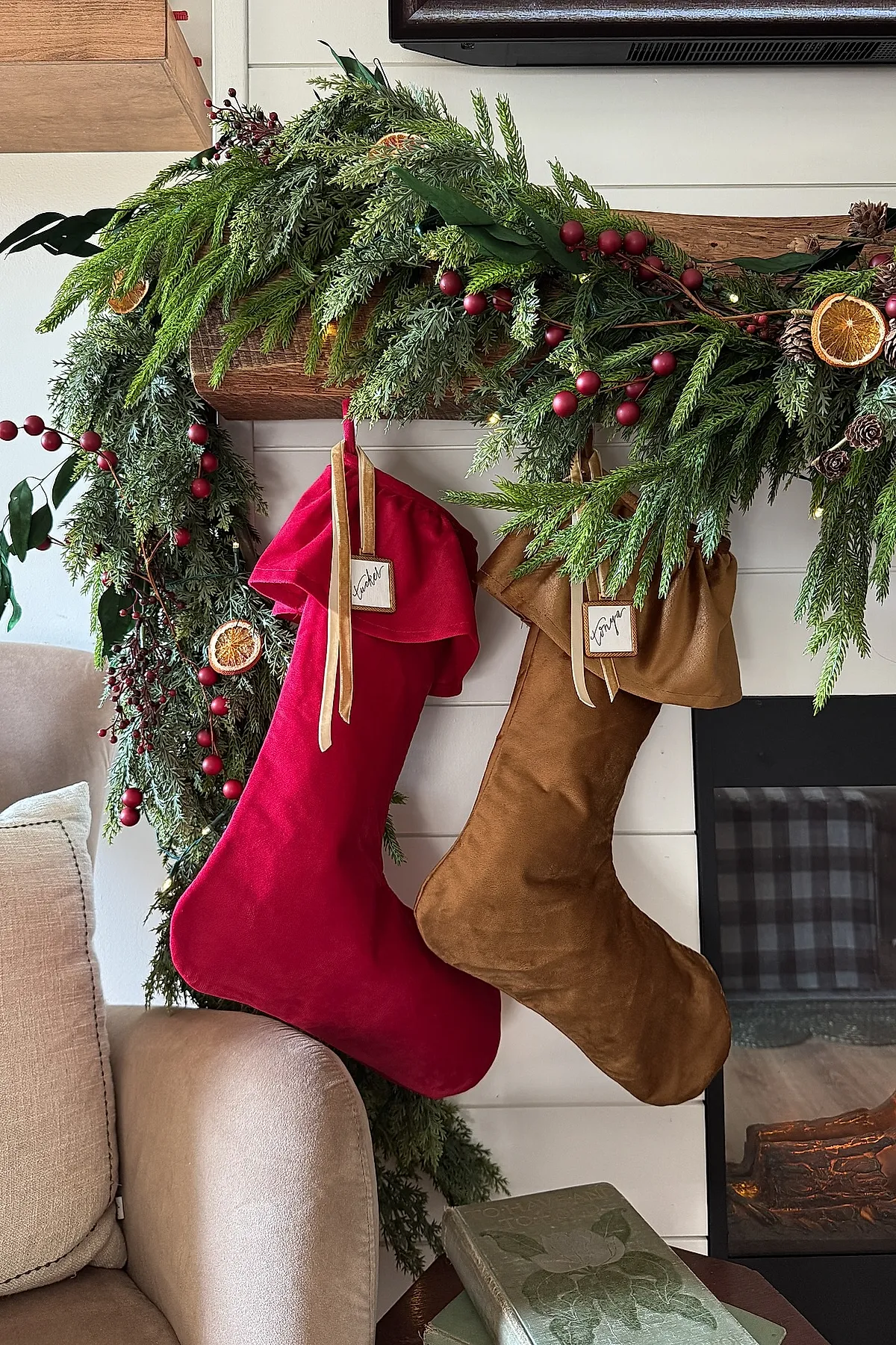 christmas mantle decorated with vibrant red and warm brown velvet stockings hanging from a wooden mantel draped in lush green garland accented with red berries and dried orange slices, above a white shiplap wall near a cozy fireplace