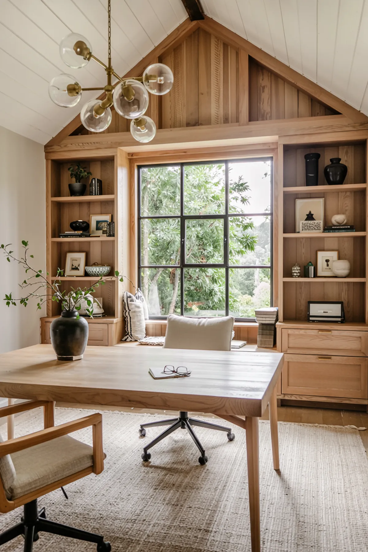 Home office with wooden shelves, large windows, modern chandelier, and cozy seating area by the window.