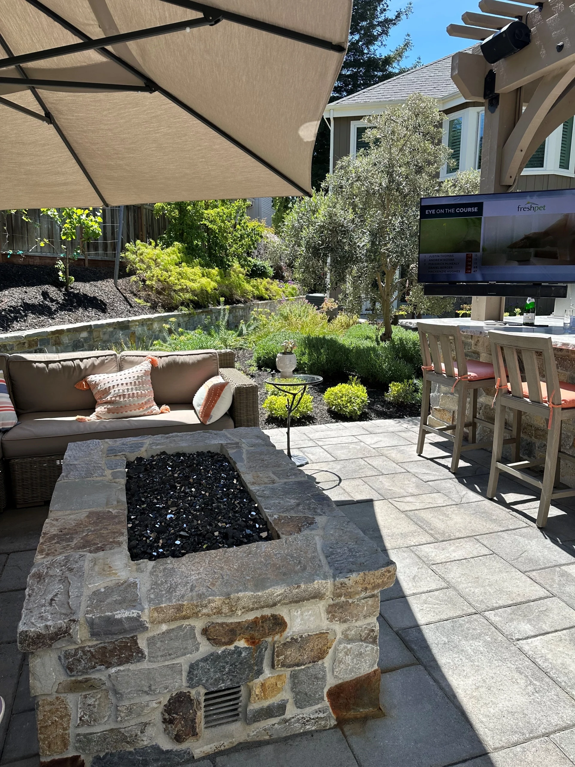 Rectangular built-in fieldstone fire pit filled with black glass rocks on a stone patio, surrounded by cushioned wicker seating, bar stools, a TV mounted under a pergola, and a large tan patio umbrella providing shade.