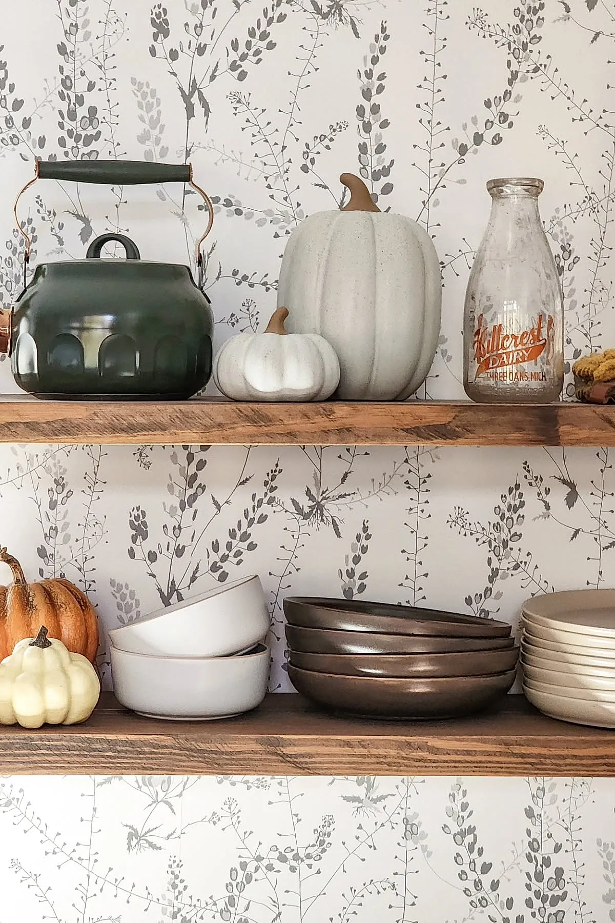 Wooden open shelves in kitchen displaying white and cream pumpkins, a dark green vintage kettle, and an old glass milk bottle against wallpaper with gray botanical prints.
