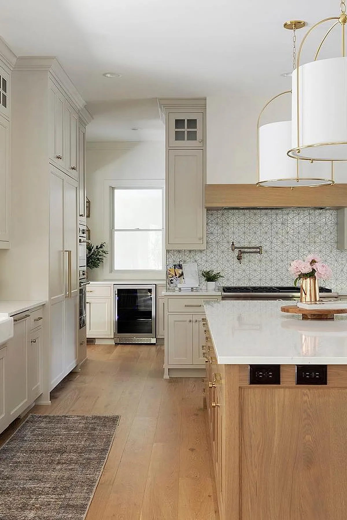 French country kitchen featuring light beige cabinets, geometric gray and white tile backsplash, large wooden island with brass hardware, white countertops, and gold-accented pendant lights.