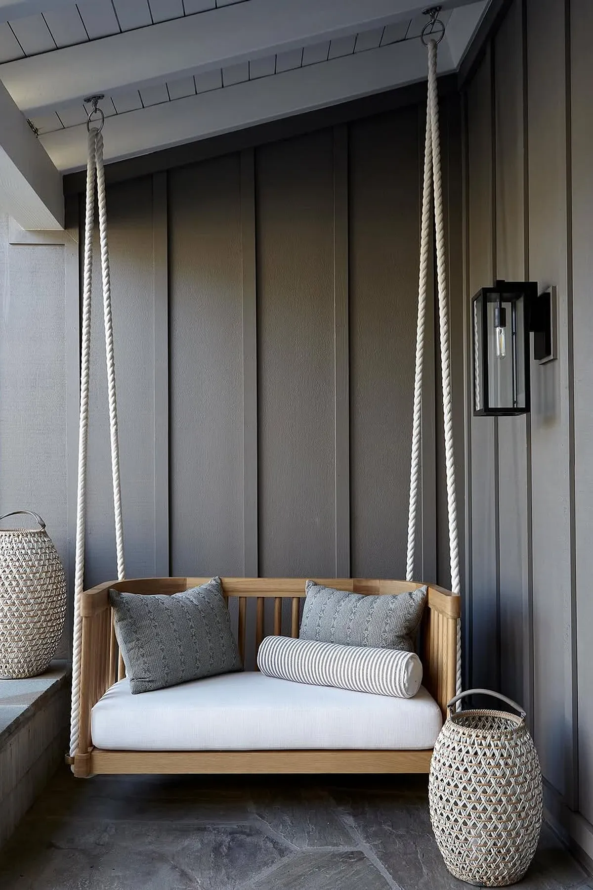 A modern porch swing in natural wood with gray pillows, suspended by thick ropes against a dark wall.