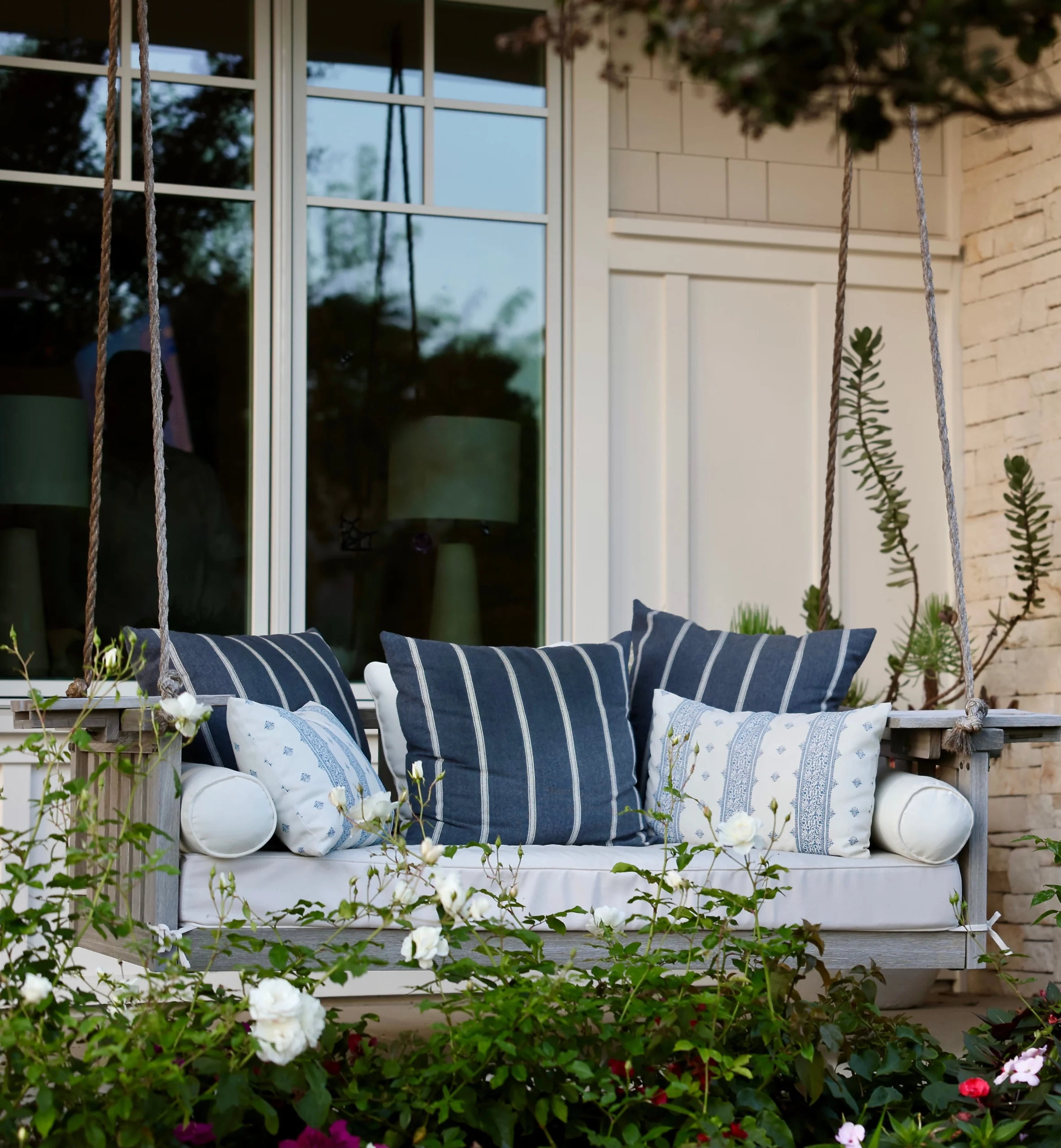 Porch swing featuring striped blue pillows, white cushions, and blooming white roses in the foreground