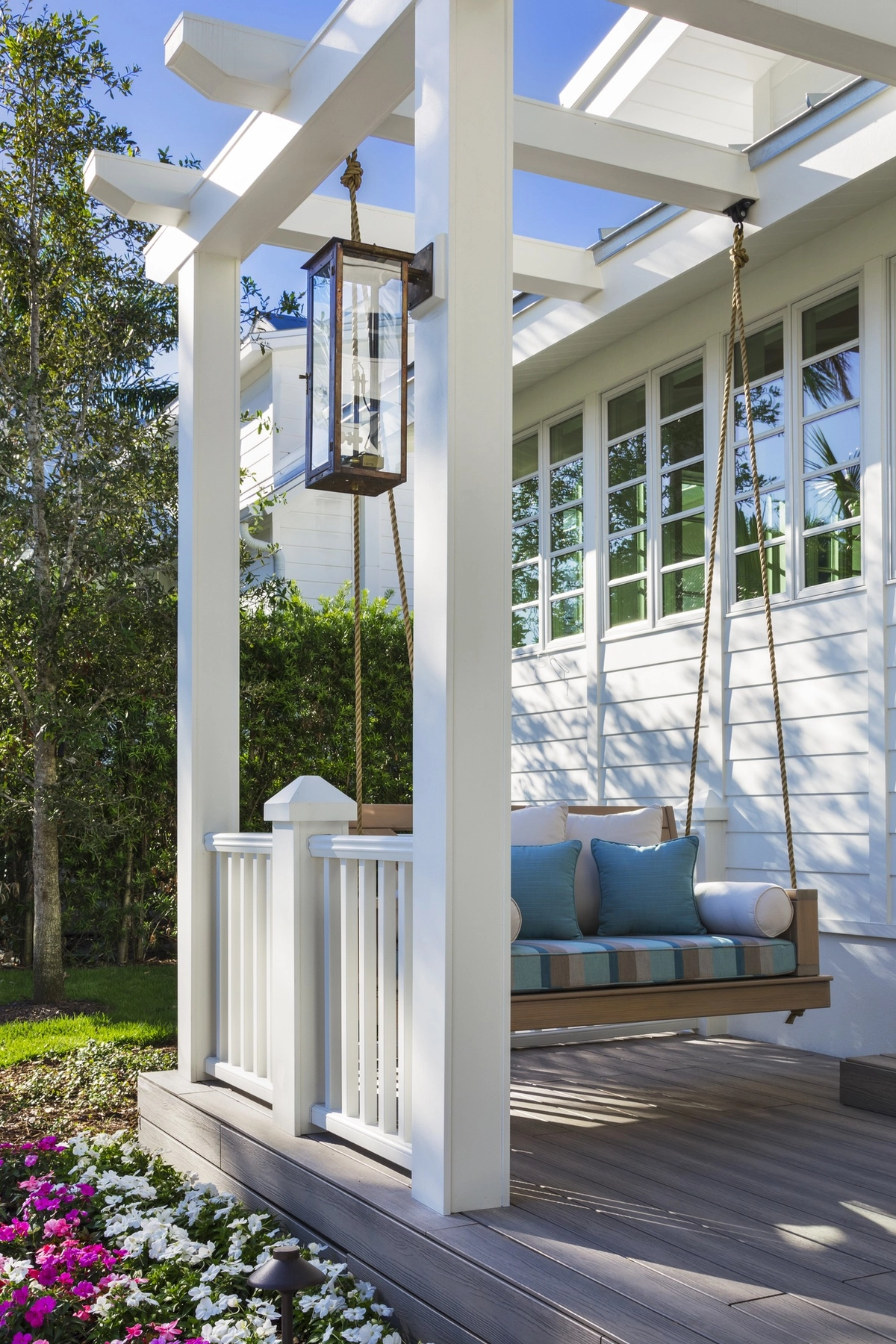 Wooden porch swing with blue striped cushions and rope hangers on a white house, surrounded by blooming flowers and greenery under a bright blue sky