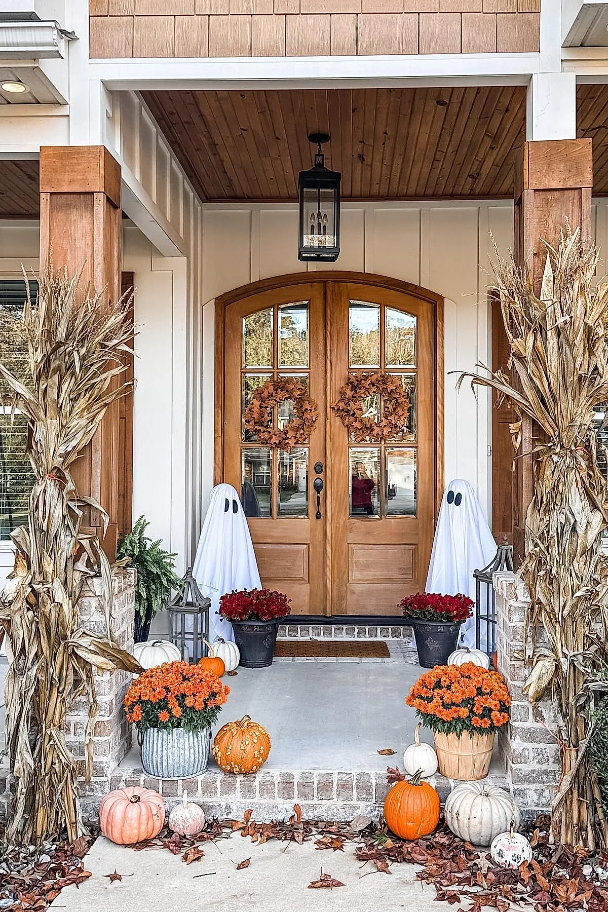 two ghost figures made from white sheets with black eyes stand beside wooden double doors decorated with autumn wreaths surrounded by orange mums orange white and pink pumpkins dried corn stalks and lanterns