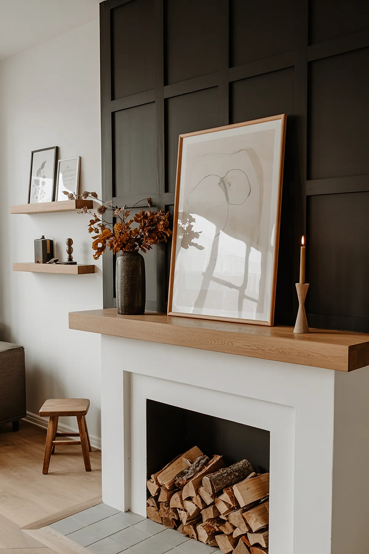 Modern fireplace with white surround and wooden mantel against dark grid-paneled wall. Mantel displays abstract framed artwork leaning against wall, tall ceramic vase holding dried brown leaves, and slim wooden candle holder with lit taper candle.