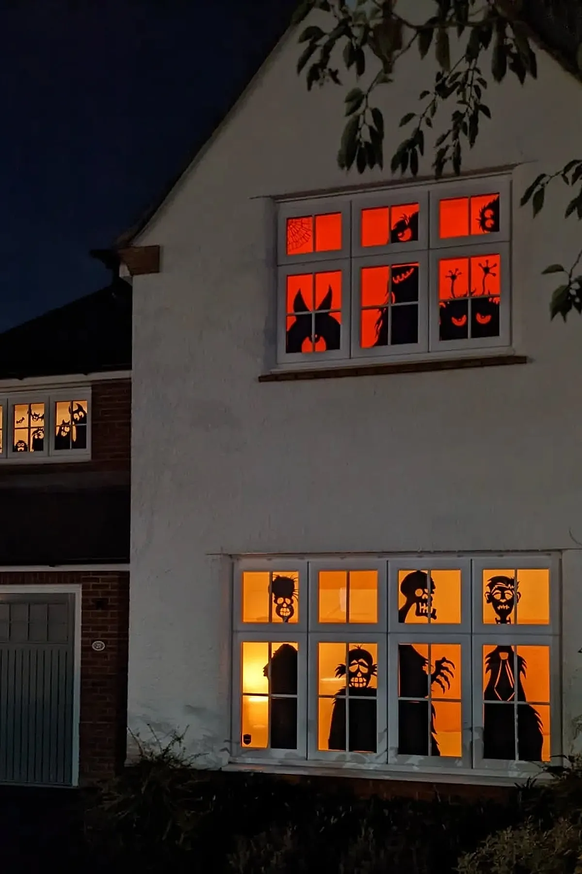 House windows glowing orange and red at night with black paper silhouettes of various cartoonish monsters, ghosts, and spooky faces arranged across multiple panes.